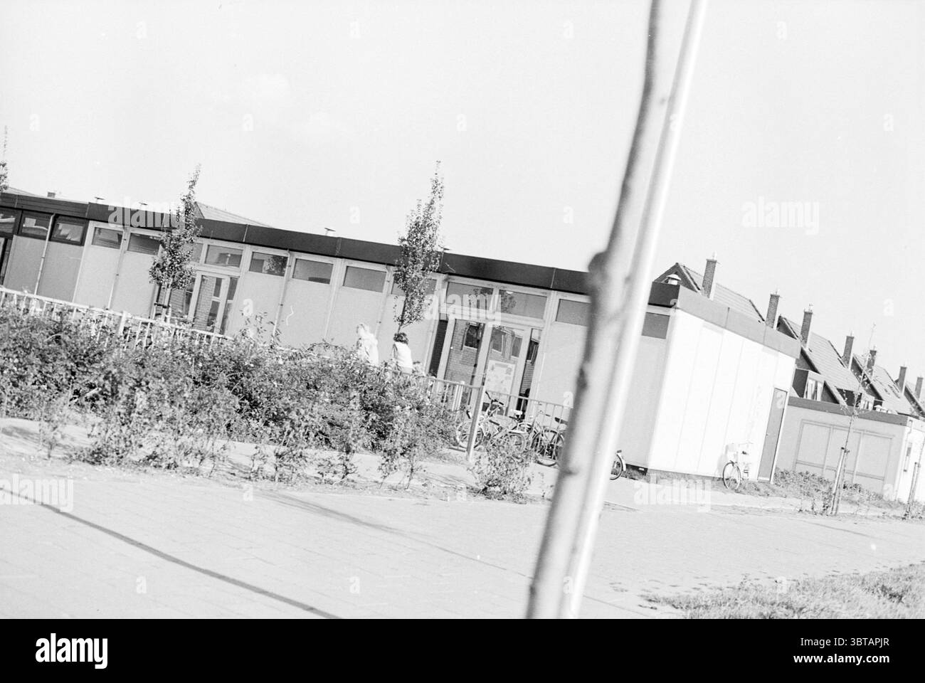 Esterno di una scuola., Whizgle News, Dutch Desk, Paesi Bassi, 1950 - 2000. Questi sono gli elementi dell'immagine. La scena cattura un ambiente urbano tranquillo caratterizzato da una miscela di spazi residenziali e comuni. In primo piano, una bassa recinzione fatta di spranghe di legno circonda una piccola macchia di verde, caratterizzata da file di arbusti ed erba che rompono la monotonia del cemento. A sinistra, due persone sono impegnate in una conversazione, posizionate leggermente dietro la recinzione. Sembrano rilassati, suggerendo un'atmosfera di interazione informale. Le loro silhouette sono sottilmente definite contro la luce Foto Stock
