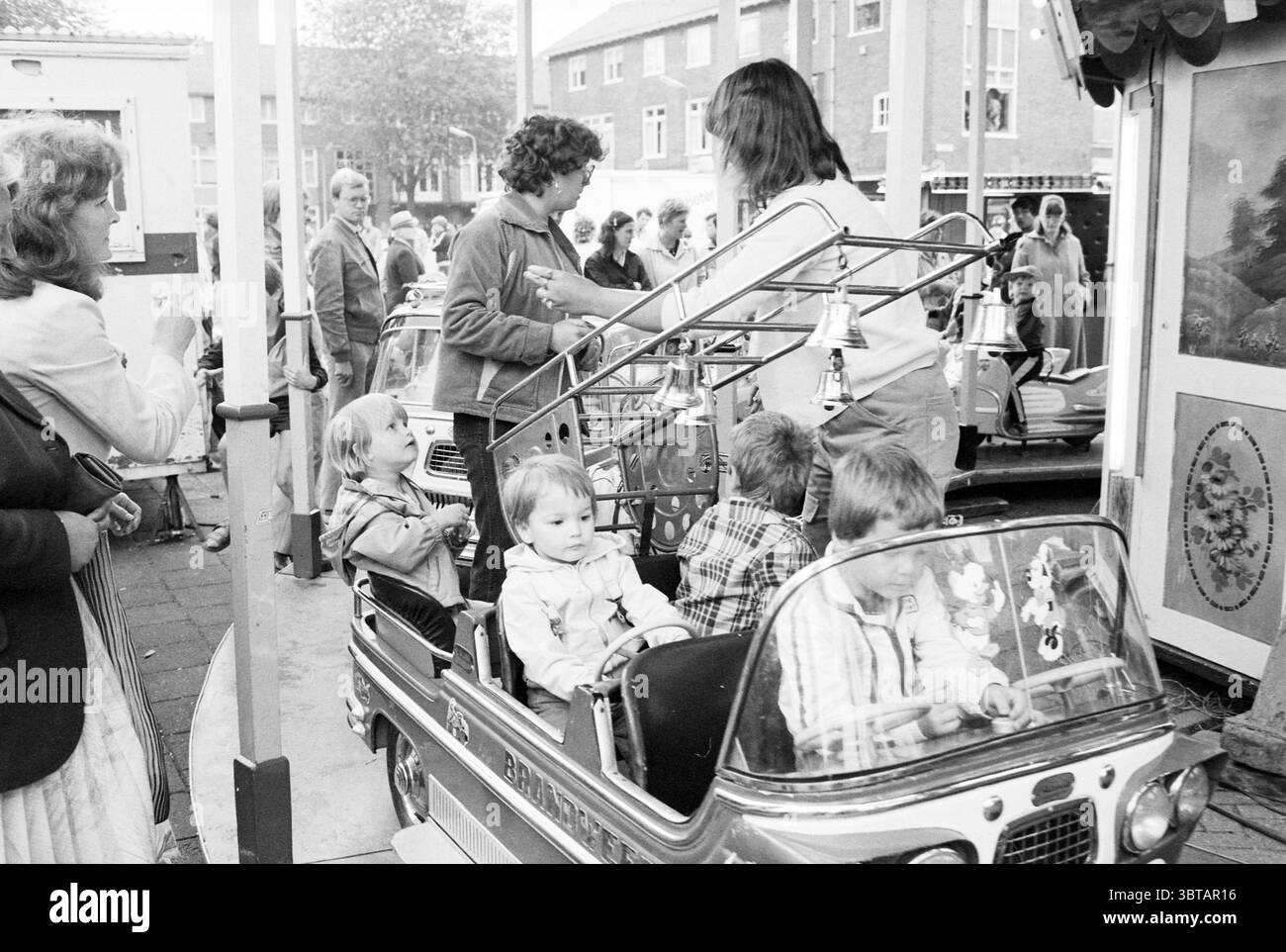 Mercato estivo IJmuiden Kennemerlaan Children on Merry-go-round IJmuiden Kennemerlaan Paesi Bassi, Whizgle News, Dutch Desk, Paesi Bassi, 1950 - 2000 il 19-06-1981. Questi sono gli argomenti nell'immagine. In una vivace scena all'aperto, una piccola area di divertimento e' piena di attivita'. L'attenzione si concentra su una serie di colorate auto vintage che i bambini stanno occupando con ansia. Le auto, lucide e metalliche, risplendono nella luce naturale soffusa, esaltandone i colori brillanti. Intorno alle giostre, diversi adulti si impegnano con i bambini, dimostrando una miscela di divertimento e cura. Un genitore si piega Foto Stock