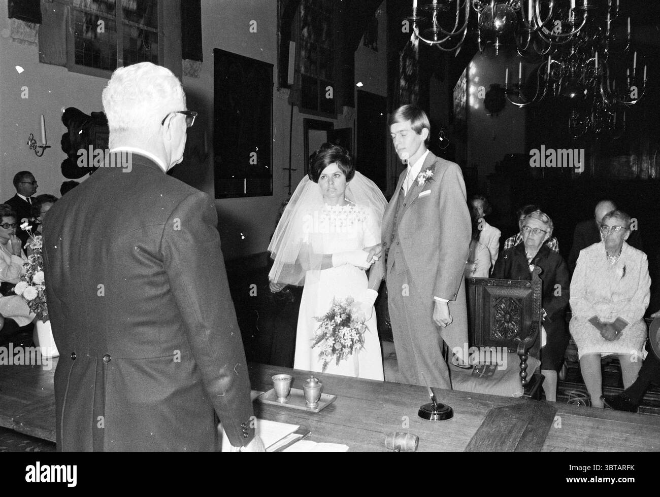 Wedding Jonkers - van Schagen Marriage, Whizgle News, Dutch Desk, Paesi Bassi, 1950 - 2000 il 16-10-1969. L'immagine contiene questi argomenti. In un ambiente solenne e intimo, una coppia si trova di fronte a un officiante, che si sta rivolgendo a loro. La donna, decorata con un classico abito bianco da sposa, indossa un velo delicato che incornicia delicatamente il suo viso. Possiede un bouquet di fiori, aggiungendo un tocco di bellezza naturale al suo gruppo. L'uomo sta accanto a lei, vestito con un abito grigio chiaro, trasuda un senso di eleganza. Le loro espressioni riflettono un mix di anticipazione e solennità, incarnandone il significato Foto Stock
