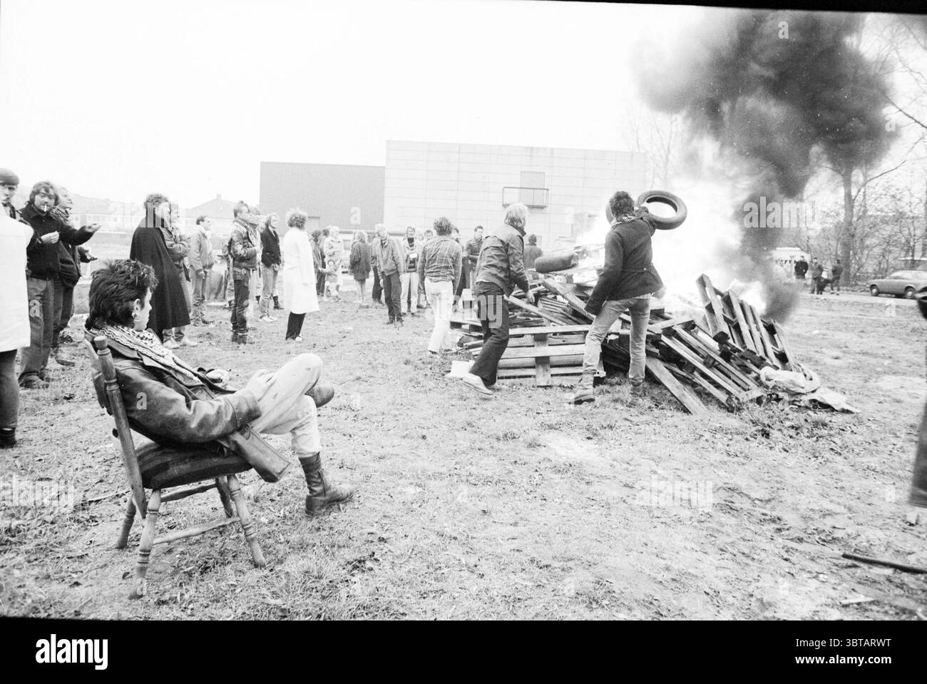 Dimostrazione contro lo sfratto del campo di carovane dimostrazione, Whizgle News, Dutch Desk, Paesi Bassi, 1950 - 2000 il 24-11-1987. Questi sono gli elementi dell'immagine. La scena raffigura un raduno all'aperto, dove un gruppo di persone è impegnato in un'attività centrata intorno a una grande pila di pallet di legno che sono in fiamme, creando un buco di fumo scuro che si innalza nell'aria. L'impostazione è piuttosto grezza, con un'area erbosa disseminata di detriti sparsi. In primo piano, una persona siede casualmente su una semplice sedia di legno, rivestita di giacca di pelle e jeans. Questo individuo appare contemplativo, wat Foto Stock