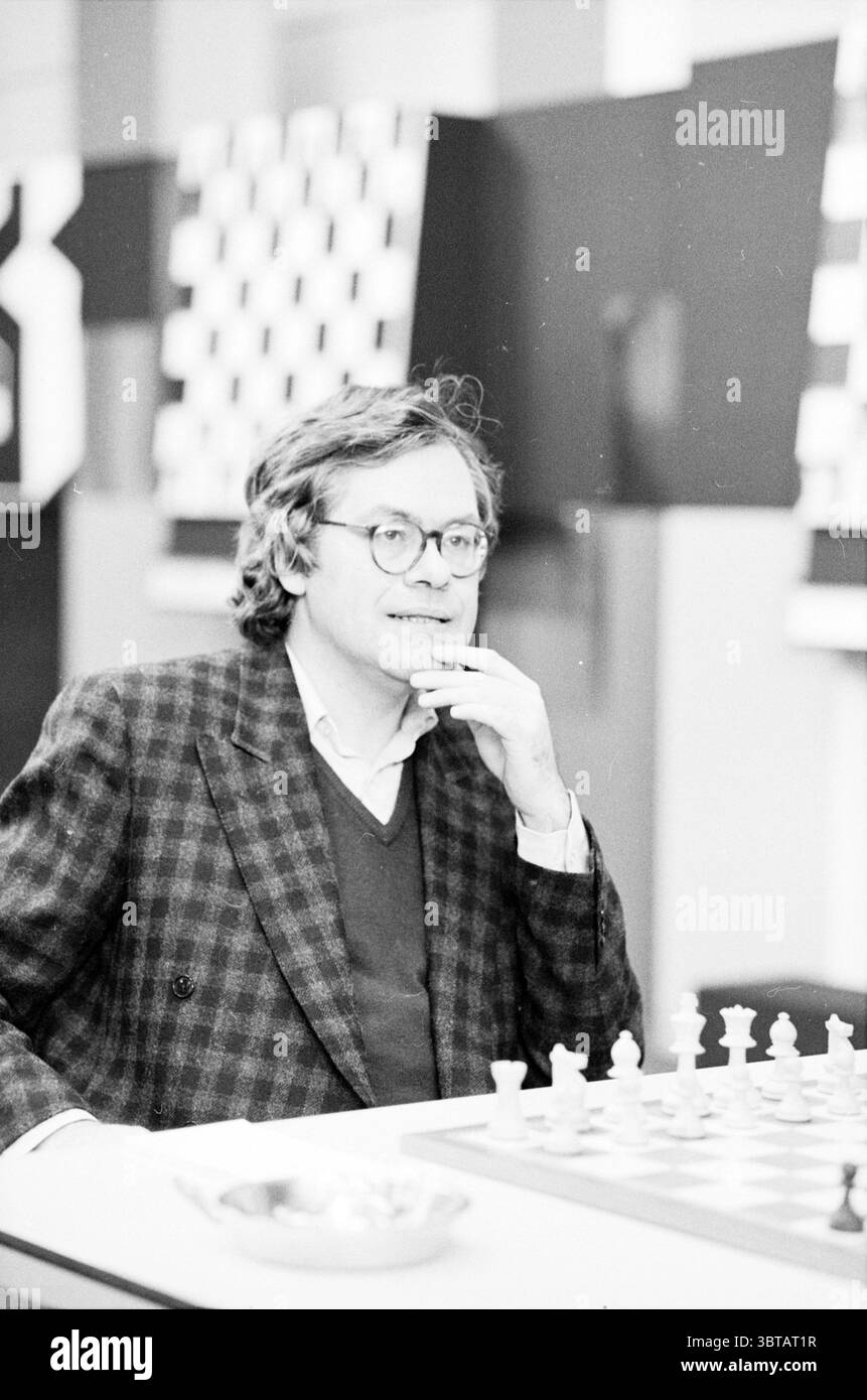 Ritratti del grande gruppo H.S.T. Blast Furnace Chess Tournament Wijk aan Zee, Whizgle News, Dutch Desk, Paesi Bassi, 1950 - 2000 il 17-01-1986. L'immagine include questi argomenti. In una scena silenziosa e monocromatica, un individuo di mezza età si siede con cura su una scacchiera, con la mano premurosa vicino al mento. Indossa un blazer dai motivi scuri che contrasta delicatamente con un maglione dalle tonalità più leggere, incarnando uno stile classico e intellettuale. Lo sfondo è decorato con tavole a scacchi, aggiungendo un senso di profondità e contesto all'impostazione. La disposizione dei pezzi su t Foto Stock
