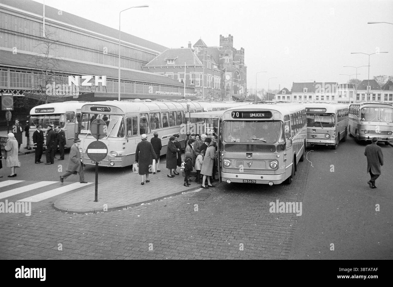 Stazione degli autobus di Haarlem, Haarlem Stationsplein Nederland., Whizgle News, Dutch Desk, Paesi Bassi, 1950 - 2000. Questi sono gli argomenti nell'immagine. La scena cattura un vivace centro di trasporti, pieno di vari autobus e gruppi di persone. I colori dominanti sono i grigi e i bianchi attenuati, indicativi di un'epoca passata, con occasionali spruzzi di colore dai vestiti dei passeggeri. A sinistra, un gruppo di autobus d'epoca, caratterizzati da forme arrotondate e accenti cromati, attendono i passeggeri. Ogni autobus ha marcature distinte, che identificano i propri percorsi. A destra, un po' di sfarzo Foto Stock
