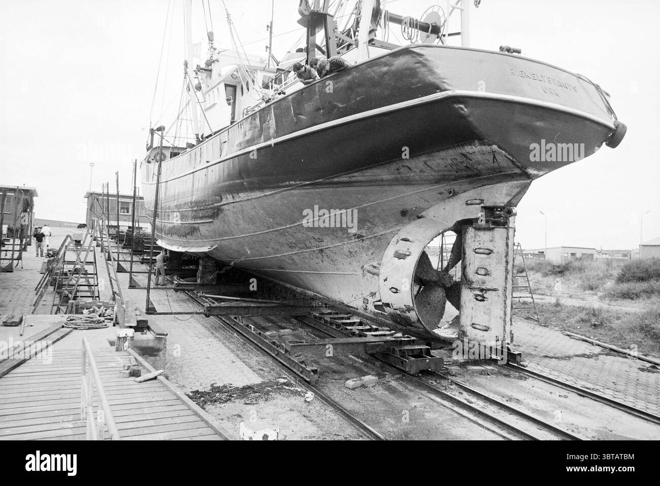 Pushboat on slipway IJmuiden IJmuiden Nederland, Whizgle News, Dutch Desk, Paesi Bassi, 1950 - 2000 il 05-08-1992. Questi argomenti sono mostrati nell'immagine. La scena cattura una grande barca da pesca che viene sollevata in modo prominente dall'acqua, poggiata su una serie di robuste rotaie in legno in un cantiere navale. La barca presenta un profondo e ricco scafo blu navy, che si erge in contrasto con la gradazione di grigi silenziosa dei suoi dintorni. Il suo ponte è adornato da una varietà di attrezzature, tra cui reti da pesca e bobine di corda, a indicare il suo uso attivo negli sforzi marittimi. Intorno al recipiente, ai macchinari e agli utensili in acciaio Foto Stock