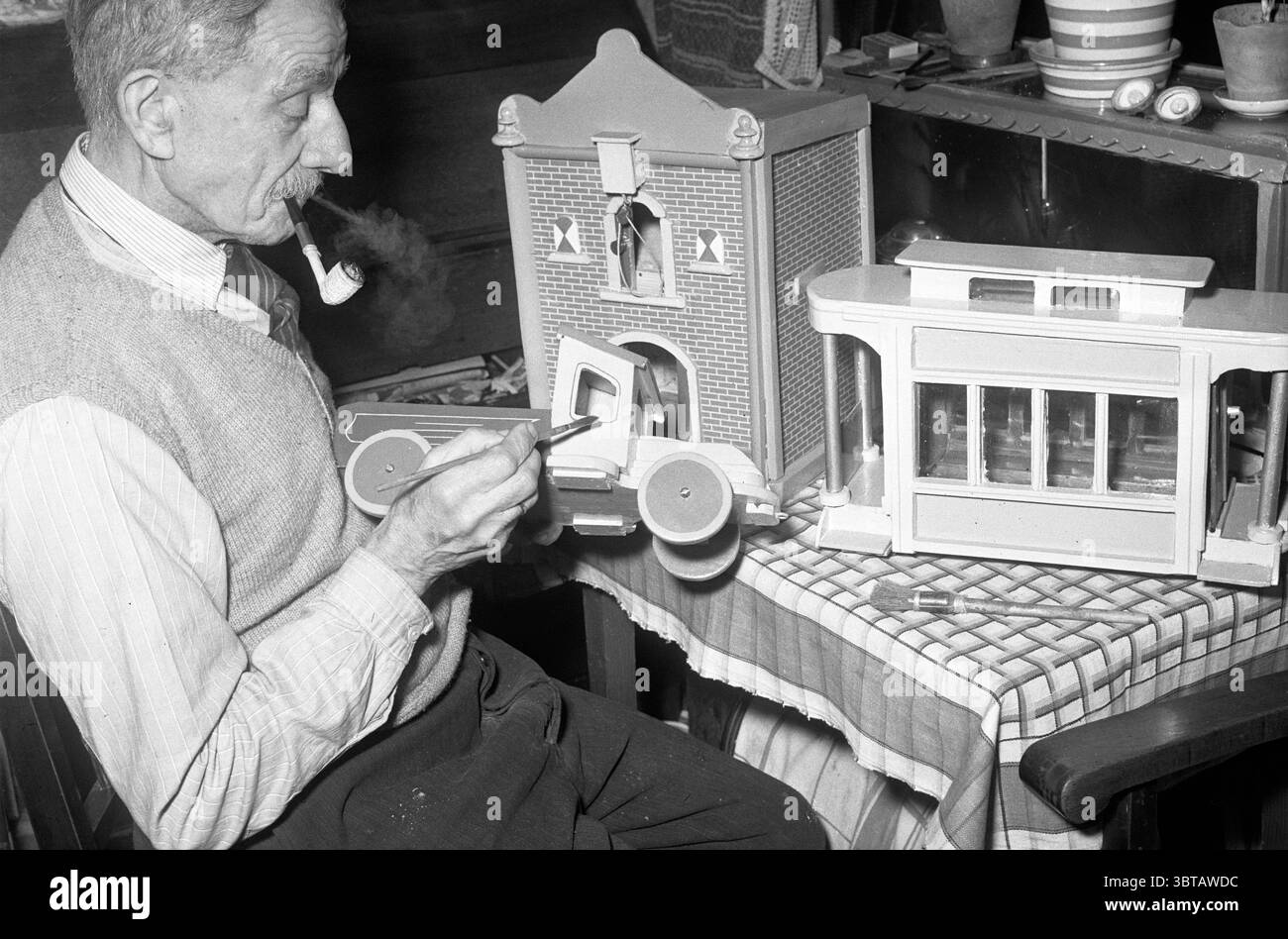 Il signor V.d. Kamp fa giocattoli di legno un camion una casa un tram, Whizgle News, Dutch Desk, Paesi Bassi, 1950 - 2000 il 21-01-1953. Questi sono gli argomenti nell'immagine. Nella scena, un uomo anziano siede su una sedia di legno, profondamente impegnato nell'atto di dipingere un camion giocattolo. Ha un aspetto distinto, caratterizzato da baffi curati e capelli grigi. Il suo abbigliamento è costituito da una camicia di colore chiaro con maniche arrotolate e un gilet di lana, suggerendo un mix di comfort e cura nel suo aspetto. L'uomo tiene in mano un pennello mentre si accoppia il furgone nell'altra. Il furgone è qui Foto Stock