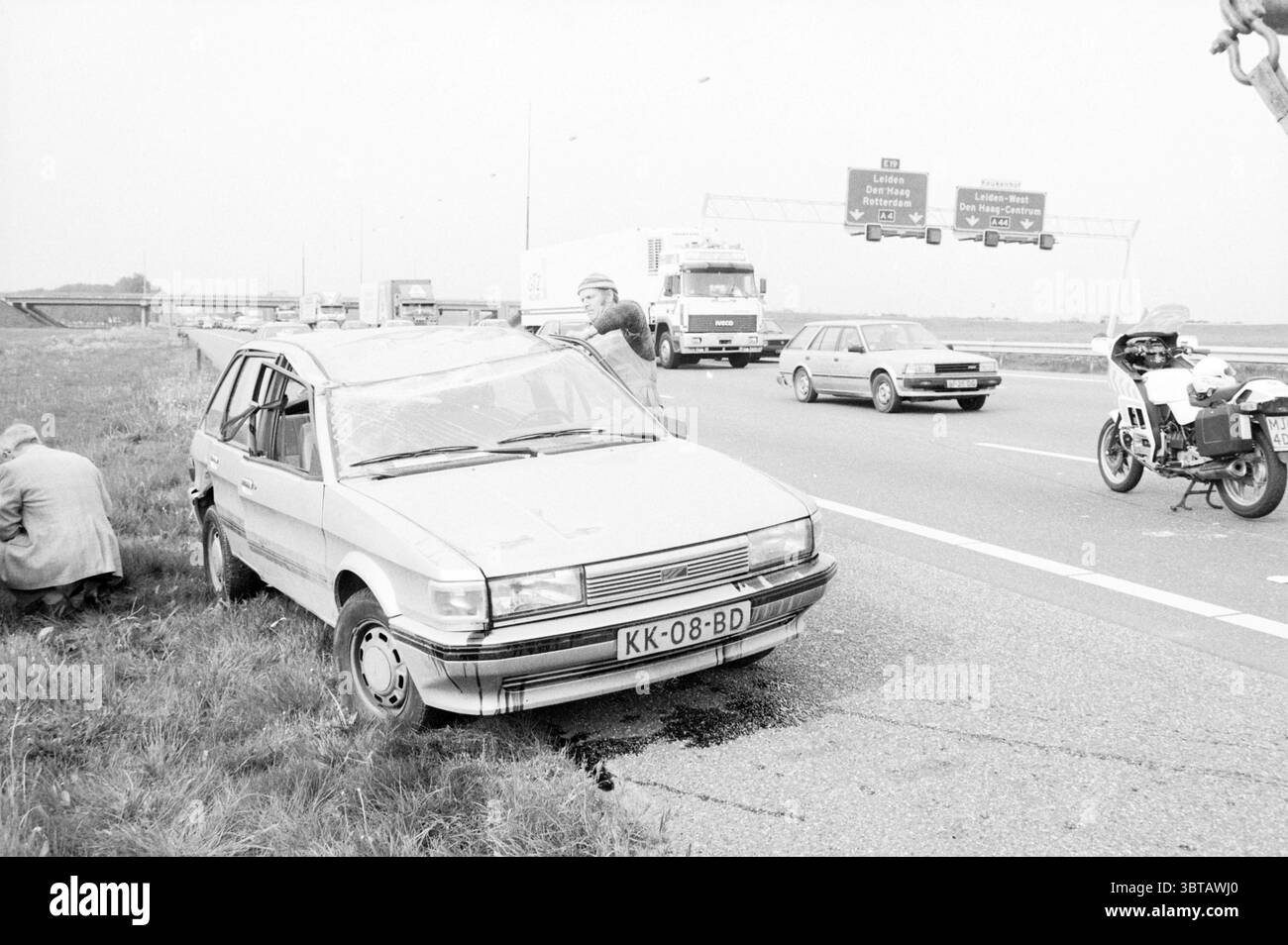 Incidente A4 vicino a Nieuw. Vennep Accidents Collisions Accidents Nieuw-Vennep Rijksweg A4, Whizgle News, Dutch Desk, Paesi Bassi, 1950 - 2000 il 11-05-1989. Questi argomenti vengono visualizzati nell'immagine. La scena raffigura una giornata grigia e ricoperta su un'autostrada trafficata. Un'auto danneggiata, verniciata con un tono basso, è parcheggiata sull'erba accanto alla strada. Il veicolo mostra segni di pericolo, con un cofano deformato e un parabrezza frantumato, a indicare che è stato coinvolto in un incidente. In primo piano, un individuo accovacciato vicino alla vettura, ispezionando potenzialmente il danno o effettuando riparazioni. Questa cifra si fonde nel cupo bancomat Foto Stock