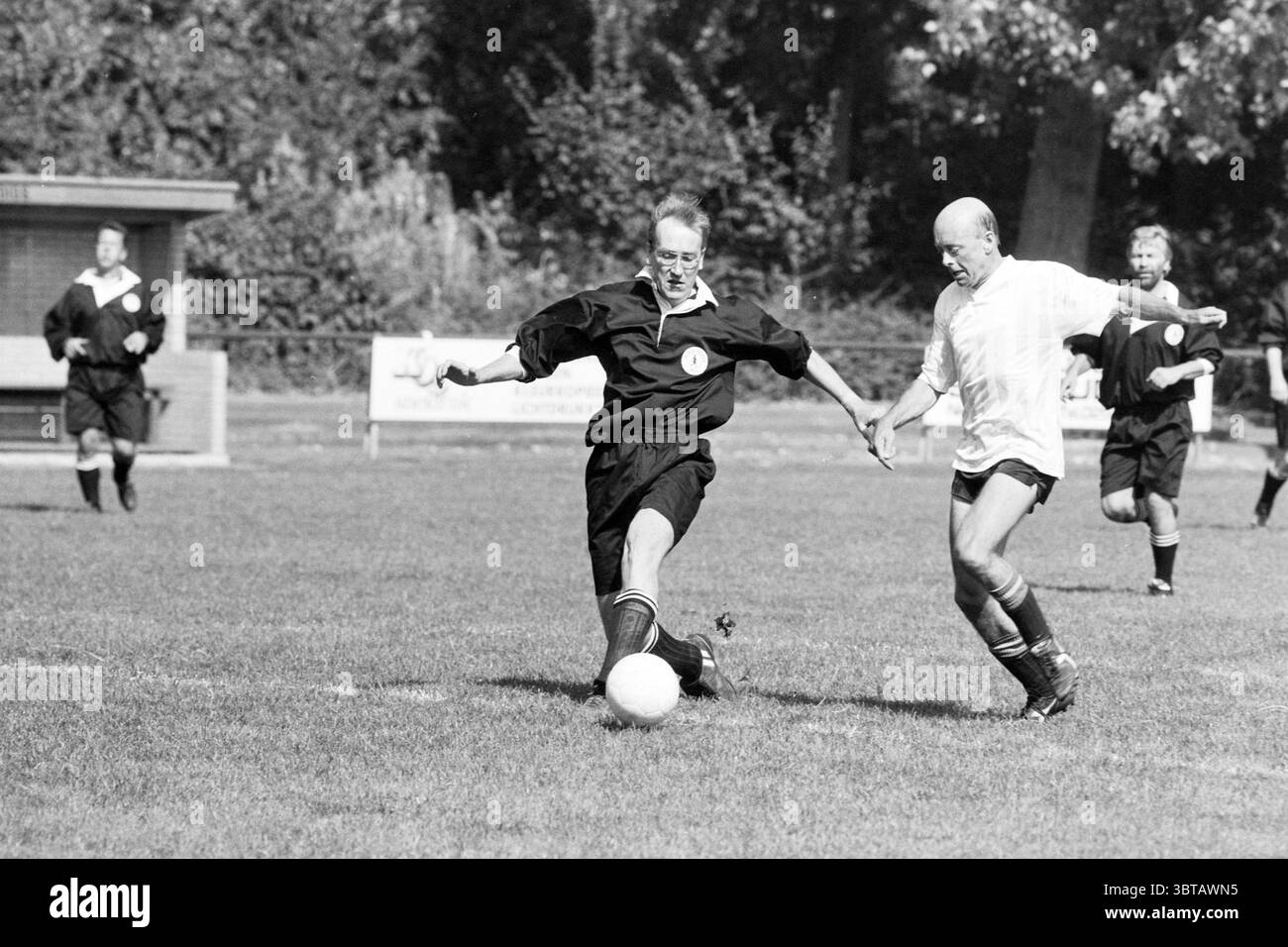Torneo Black Devils Nieuw Vennep Nieuw-Vennep, Whizgle News, Dutch Desk, Paesi Bassi, 1950 - 2000 il 30-08-1991. L'immagine include questi argomenti. In una scena dinamica su un campo verde e ben tenuto, due atleti si impegnano in un intenso momento di gioco durante una partita di calcio. In primo piano è presente un arbitro, vestito con una classica uniforme nera con un distintivo, che colpisce abilmente la palla con il piede destro, facendola avanzare. I suoi occhiali riflettono un senso di concentrazione e di autorità, mentre la sua postura trasmette determinazione e attenzione. Opponendosi a lui è un giocatore con una maglia bianca, Foto Stock