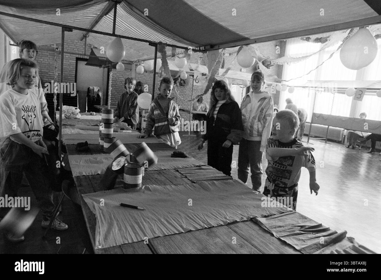 Il giorno della Regina. The Meerkoet Lisserbroek Queen's Day Lisserbroek, Whizgle News, Dutch Desk, Paesi Bassi, 1950 - 2000 il 30-04-1990. Questi argomenti vengono visualizzati nell'immagine. In un ambiente interno spazioso, un gruppo di bambini è impegnato in un'attività vivace. La sala è decorata con decorazioni colorate che suggeriscono un'atmosfera festosa, probabilmente per una celebrazione o un evento. In primo piano, viene allestito un tavolo fatto di tavole di legno, dove alcuni bambini sono riuniti, mostrando entusiasmo mentre partecipano a un gioco. Un bambino che indossa una t-shirt di colore chiaro con grafica a fumetti, è in movimento, trowin Foto Stock