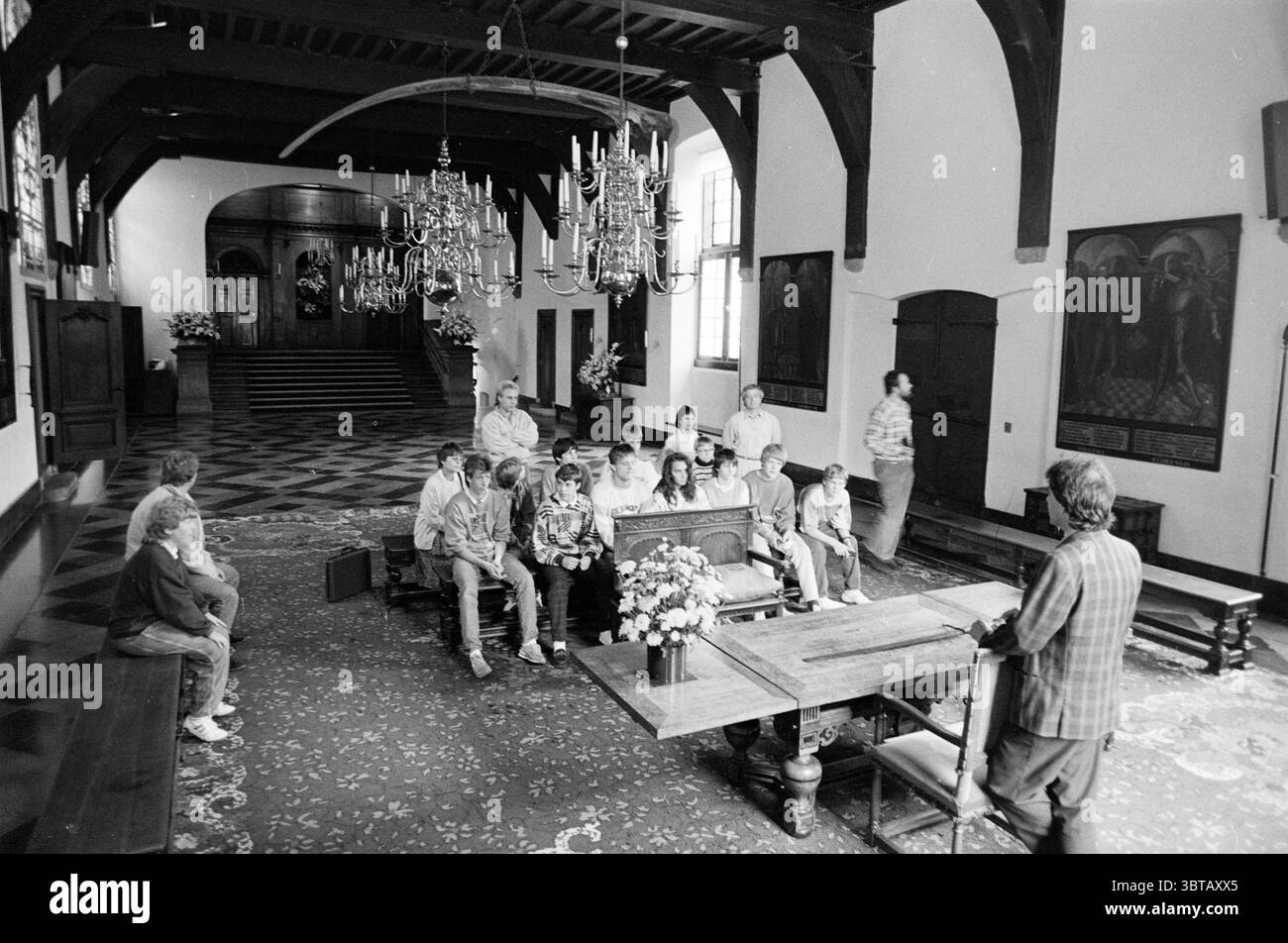 Gruppo di studenti di Osnabrück al municipio gruppi studenti Haarlem Grote Markt Paesi Bassi, Whizgle News, Dutch Desk, Paesi Bassi, 1950 - 2000 il 19-09-1988. L'immagine contiene questi argomenti. La scena presenta un interno spazioso e ricco di storia caratterizzato da alte travi in legno che attraversano un soffitto, contribuendo a un senso di grandezza. Il pavimento e' adornato con piastrelle con motivi intricati e un grande tappeto ornato che aggiunge calore alla camera. Riuniti in un semicerchio vi sono un gruppo eterogeneo di persone sedute su panchine e sedie, rivolti con attenzione verso un altoparlante davanti. L'indo Foto Stock
