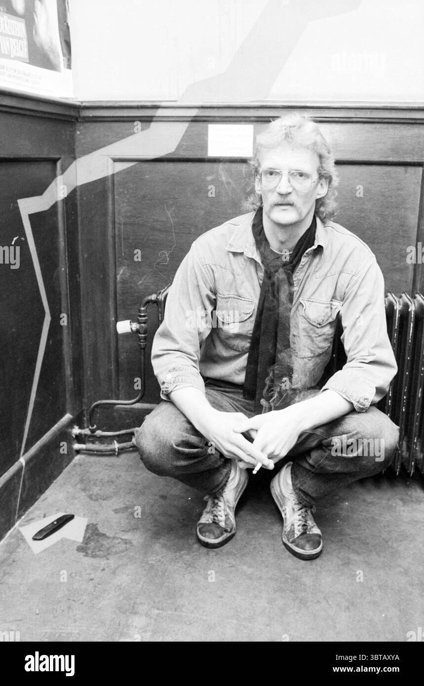 Wall drawing café Plok Drawings drawings, Whizgle News, Dutch Desk, Paesi Bassi, 1950 - 2000 il 08-04-1986. Questi argomenti sono mostrati nell'immagine. In uno spazio interno scarsamente illuminato, un uomo è posizionato in una posa accovacciata sul pavimento. Il suo abbigliamento è composto da una camicia in denim azzurro abbinata a jeans leggermente affaticati, che suggerisce un'atmosfera casual o artistica. Una sciarpa nera legata al collo aggiunge un tocco di contrasto e stile al suo outfit. Ha i capelli taglienti e a spalla e sfoggia baffi spessi, contribuendo a creare un'estetica bohémien o vintage. Con un'espressione premurosa Foto Stock