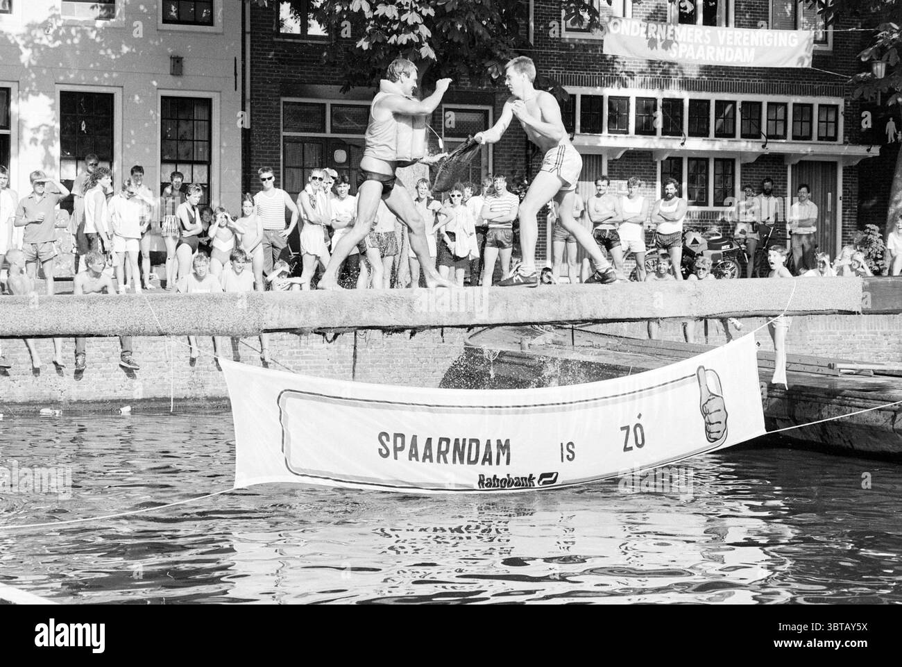 Ponderslaan in Spaarndam giochi e giocattoli Spaarndam, Whizgle News, Dutch Desk, Paesi Bassi, 1950 - 2000 il 28-06-1986. Questi argomenti sono mostrati nell'immagine. La scena è vivace e dinamica, catturando l'energia di un evento all'aperto. Due persone, che indossano pantaloncini corti, sono posizionate in un coinvolgente tussle su una piattaforma stretta che si estende su un corpo d'acqua. Le loro espressioni trasmettono determinazione ed eccitazione, mentre sembrano cercare di superare l'un l'altro in una competizione giocosa. Intorno a loro, una folla diversificata di spettatori guarda con attenzione, le loro espressioni variano da divertimento a divertimento Foto Stock