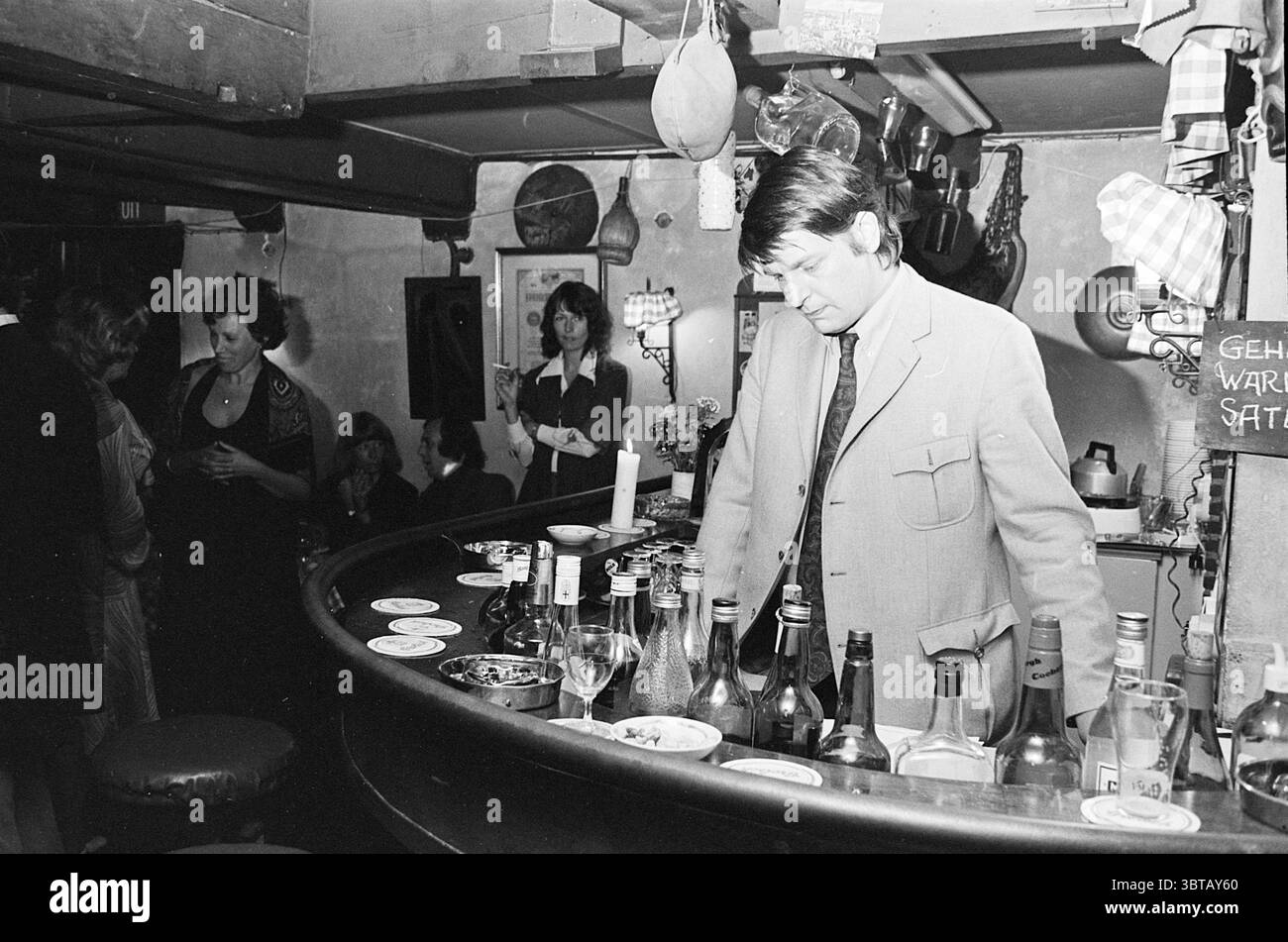 Serata di festa del club di tiro, Whizgle News, Dutch Desk, Paesi Bassi, 1950 - 2000. Questi argomenti sono mostrati nell'immagine. La scena si sviluppa in un bar accogliente e poco illuminato pieno di un'atmosfera calda e intima. Il bar stesso è circolare e luminoso, con una serie di bottiglie che rivestono il retro, che mostrano vari liquori e bevande. Il soffice bagliore dell'illuminazione ambientale proietta ombre delicate, migliorando l'atmosfera generale di relax e cameratismo. Dietro il bar, un uomo, vestito con un abito su misura, si piega leggermente in avanti, apparentemente immerso nel pensiero o magari sorseggiando un drink. I suoi capelli sono perfettamente co Foto Stock