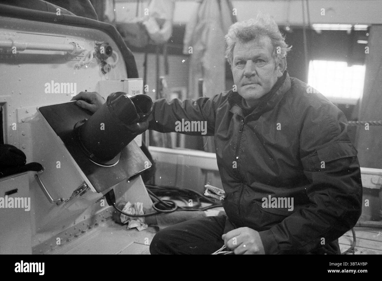 Radar per KNZHRM IJmuiden Noord Zuid Hollandse Vervoersmaatschappij N.V. NZ IJmuiden Paesi Bassi, Whizgle News, Dutch Desk, Paesi Bassi, 1950 - 2000 il 22-02-1983. Questi argomenti sono mostrati nell'immagine. La scena cattura un individuo duro seduto su un'imbarcazione, trasuda un'aria di determinazione ed esperienza. L'uomo ha un volto infranto, caratterizzato da linee prominenti che raccontano storie di una vita trascorsa all'aperto. I suoi capelli sono arricciati, suggerendo un comportamento casual ma impegnativo. È vestito con una giacca pesante e scura che contrasta con l'ambiente più leggero, suggerendo la praticità Foto Stock
