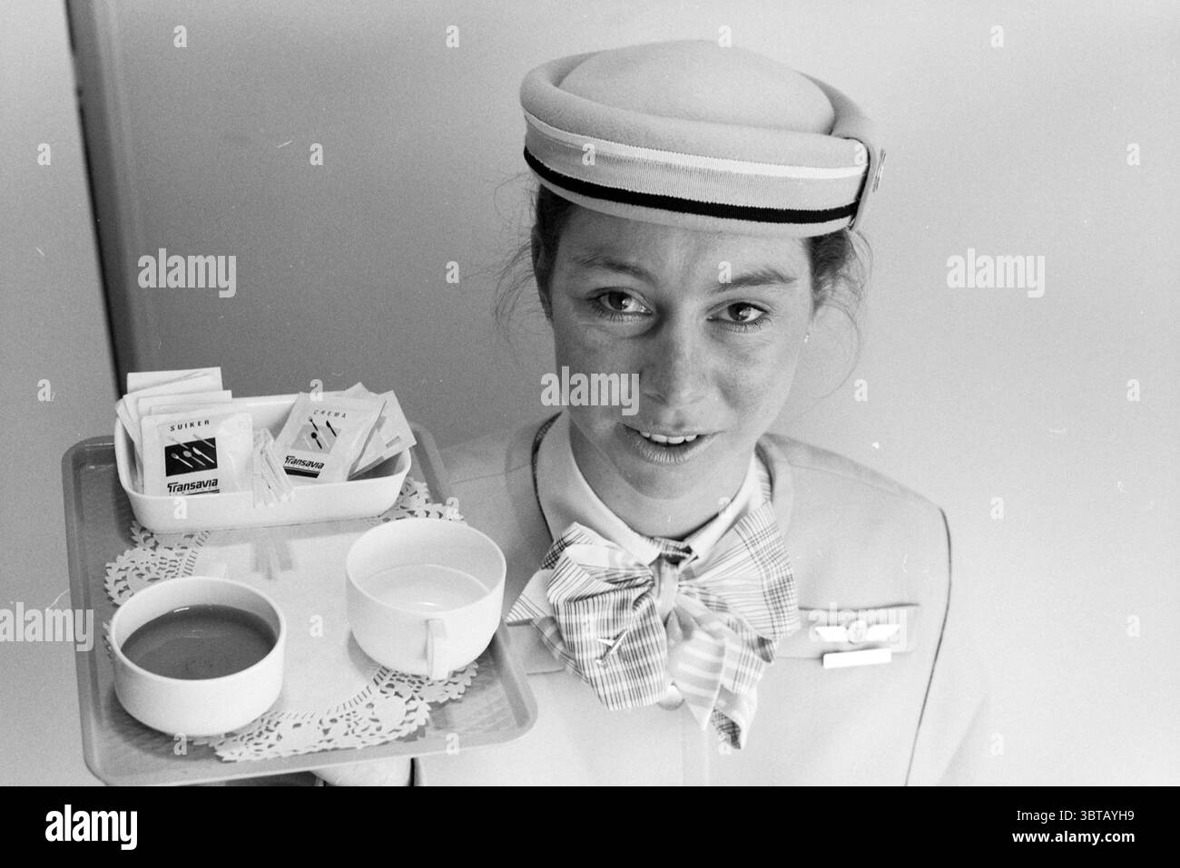 Schipholwerk Transavia assistente di volo Schiphol, Whizgle News, Dutch Desk, Paesi Bassi, 1950 - 2000 il 25-08-1991. Questi sono gli elementi dell'immagine. In questa scena, una donna che indossa un'uniforme di colore chiaro con un berretto prominente sta in piedi con sicurezza, presentando un vassoio davanti a lei. L'uniforme presenta un design ordinato e strutturato che suggerisce professionalità. Il berretto è decorato con una fascia scura che aggiunge un tocco di eleganza. Il vassoio che tiene è accuratamente sistemato, con una piccola tazza bianca riempita di una bevanda, possibilmente tè o caffè, accanto a un piatto contenente vari pacchetti di sugo Foto Stock