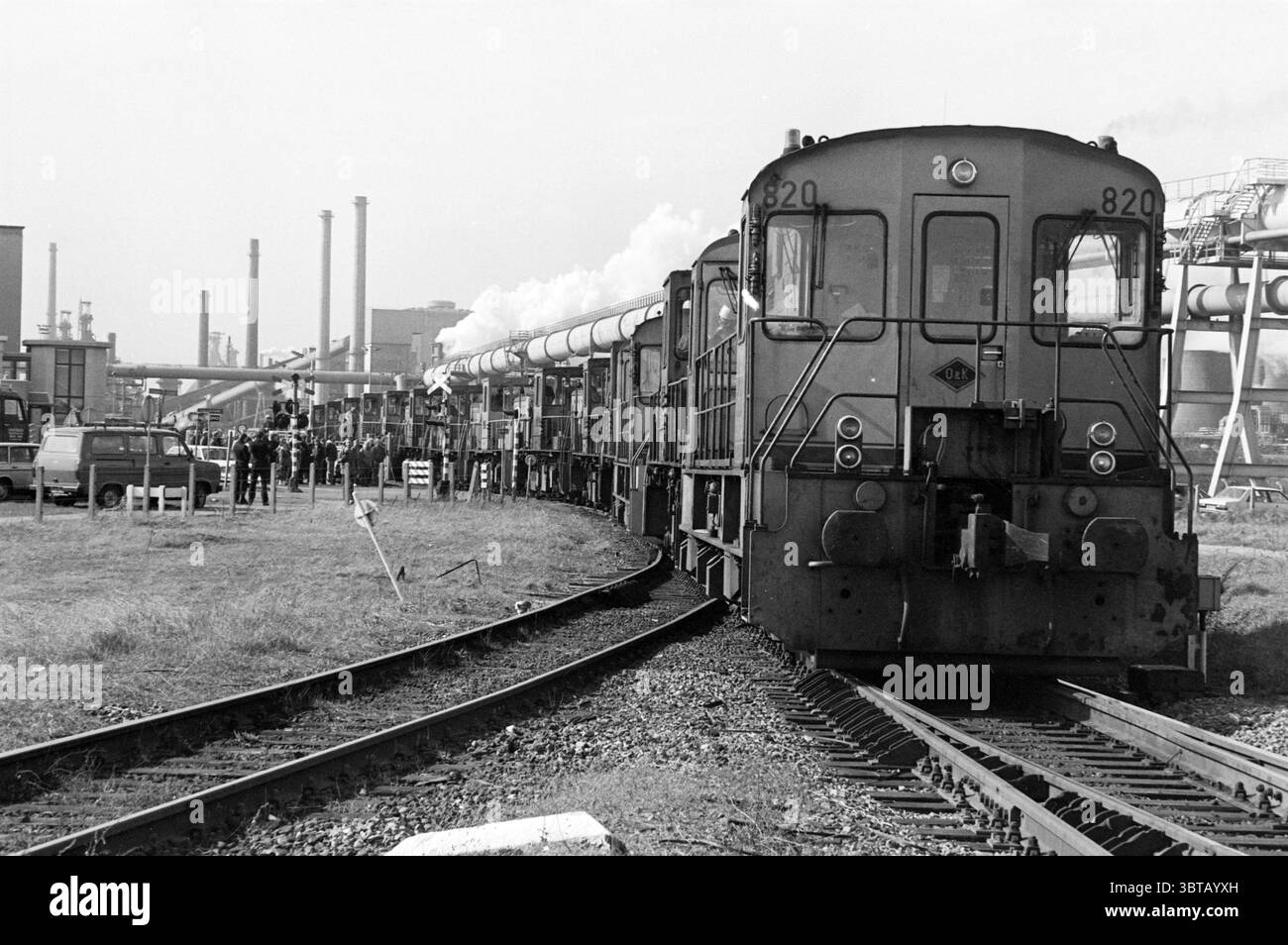Locomotive accoppiate sito Hoogovens Hoogovens, Whizgle News, Dutch Desk, Paesi Bassi, 1950 - 2000 il 01-04-1985. L'immagine contiene questi argomenti. La scena cattura un ambiente industriale vivace, caratterizzato da una coppia di treni che navigano lentamente su binari curvi. Il primo piano presenta una locomotiva diesel, caratterizzata da una struttura robusta e angolare e vernice scura, con il numero "820" ben visibile sulla parte anteriore. Intorno al treno, diverse carrozze si estendono in lontananza, alcune rilasciano delicati colpi di vapore. Sullo sfondo, le pile di fumo si innalzano contro lo skyline, Foto Stock