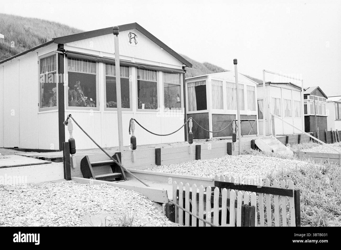 Casa sulla spiaggia omicidio Bloemendaal spiaggia e spiagge Bloemendaal, Whizgle News, Dutch Desk, Paesi Bassi, 1950 - 2000 il 21-06-1982. Questi argomenti sono mostrati nell'immagine. La scena presenta una fila di capanne sulla spiaggia, ognuna delle quali mostra un carattere unico e condivide uno stile architettonico simile. Le capanne sono prevalentemente bianche, con alcune caratterizzate da finiture più scure, e ognuna ha una linea del tetto distinta. La parte anteriore delle capanne presenta grandi finestre che riflettono la luce soffusa, offrendo scorci di interni che suggeriscono un soggiorno informale sulla spiaggia, con tocchi di mobili e decorazioni visibili all'interno. I setti Foto Stock