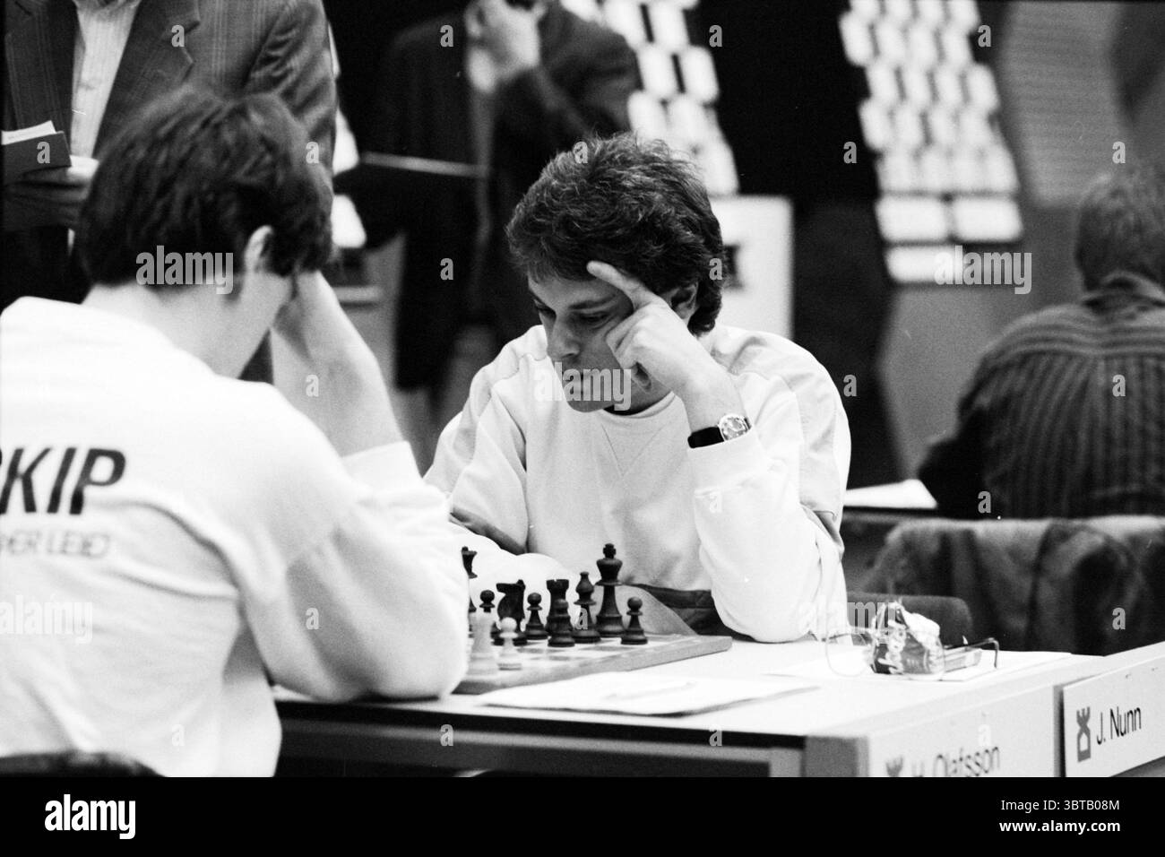 Panoramica di decathlons H.S.T. [Blastovens Chess Tournament], Whizgle News, Dutch Desk, Paesi Bassi, 1950 - 2000 il 31-01-1991. Questi sono gli elementi dell'immagine. La scena cattura una partita di scacchi tesa che si svolge in un torneo. Due giocatori sono seduti uno di fronte all'altro, concentrati sulla scacchiera che si trova tra di loro. Un giocatore, vestito con una camicia a maniche lunghe di colore chiaro, ha la mano alzata sul tempio, incarnando concentrazione e attenzione. Il suo avversario dai capelli più scuri, che indossa un abbigliamento più scuro, si appoggia leggermente, gli occhi fissati sul tabellone, contemplando il suo Foto Stock