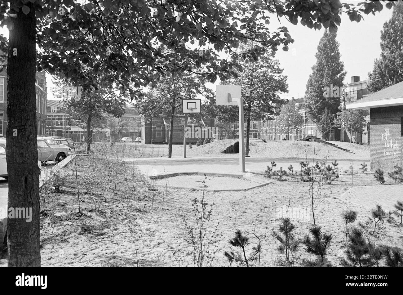 Panoramica Leidscheplein Haarlem Overviews Square Haarlem Leidseplein Paesi Bassi, Whizgle News, Dutch Desk, Paesi Bassi, 1950 - 2000 il 18-05-1976. L'immagine include questi argomenti. La scena raffigura una tranquilla area esterna, caratterizzata da una miscela di elementi naturali e urbani. In primo piano, un campo da basket rettangolare è caratterizzato da una superficie liscia e sabbiosa, con il suo cerchio ben visibile sullo sfondo di alberi ed edifici. A sinistra, il robusto tronco di un albero si erge a sentinella, che lascia un verde vibrante in contrasto con i toni tenui del terreno sabbioso, il che suggerisce un Foto Stock