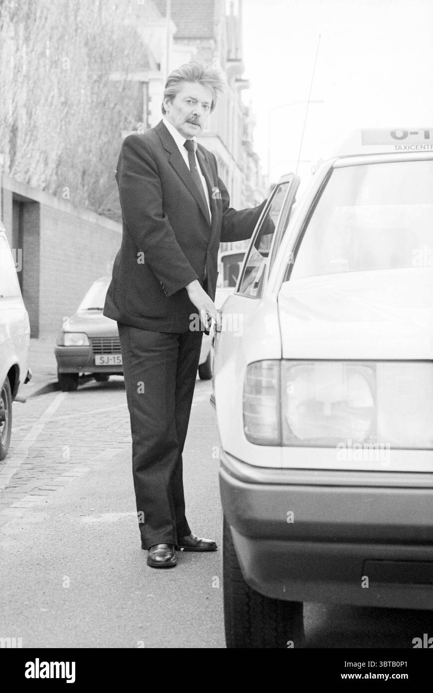 Autista in Suit o-Tax Haarlem taxi Haarlem Paesi Bassi, Whizgle News, Dutch Desk, Paesi Bassi, 1950 - 2000 il 15-02-1988. L'immagine include questi argomenti. In un ambiente urbano monocromatico, un uomo ben vestito si trova accanto a un taxi, con la mano appoggiata sulla porta. Trasuda un'aria di raffinatezza, sfoggia un abito sartoriale e scarpe raffinate che risplendono delicatamente nella luce soffusa. I suoi capelli sono ben modellati, e porta un'espressione premurosa, suggerendo che sta contemplando la sua prossima mossa. L'ambiente circostante è caratterizzato da una strada tranquilla fiancheggiata da veicoli parcheggiati e dalle loro superfici verniciate Foto Stock