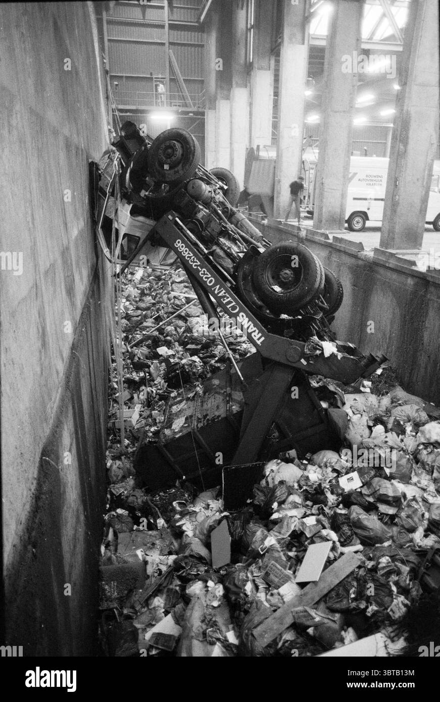 Camion della spazzatura nello scivolo, Whizgle News, Dutch Desk, Paesi Bassi, 1950 - 2000 il 12-08-1991. Questi argomenti vengono visualizzati nell'immagine. La scena raffigura un ambiente caotico e disordinato all'interno di una struttura industriale, illuminato da luci soffuse che proiettano ombre dure in tutta l'area. In primo piano, un veicolo per la raccolta dei rifiuti giace su un lato, inclinato precariamente contro una parete di cemento. Gli pneumatici del carrello sono visibili, con la gomma usurata in contrasto con il cemento grigio freddo. Intorno al carrello è presente un cumulo di detriti, costituito da carta deformata, materiali di imballaggio scartati e varie t Foto Stock