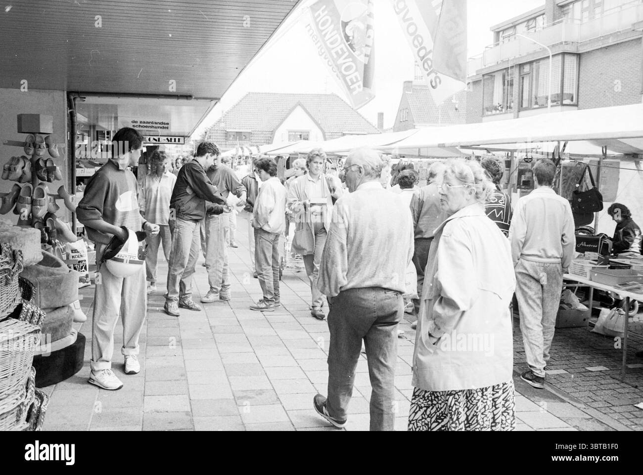 Braderie Heemskerk Braderie Heemskerk, Whizgle News, Dutch Desk, Paesi Bassi, 1950 - 2000 il 29-08-1987. Questi argomenti vengono visualizzati nell'immagine. La scena si snoda in una vivace area del mercato, caratterizzata da un'atmosfera vivace. Gruppi di persone popolano lo spazio, impegnandosi in conversazioni e sfogliando le bancarelle. È rappresentata una gamma di età, dagli anziani agli adulti più giovani, che trasmettono una presenza comunitaria diversificata. Le bancarelle fiancheggiano il sentiero, ricoperte da tettoie colorate che forniscono ombra dalla luce dall'alto. I colori delle tende da sole variano, aggiungendo un vibrante contrasto visivo al neutro a. Foto Stock