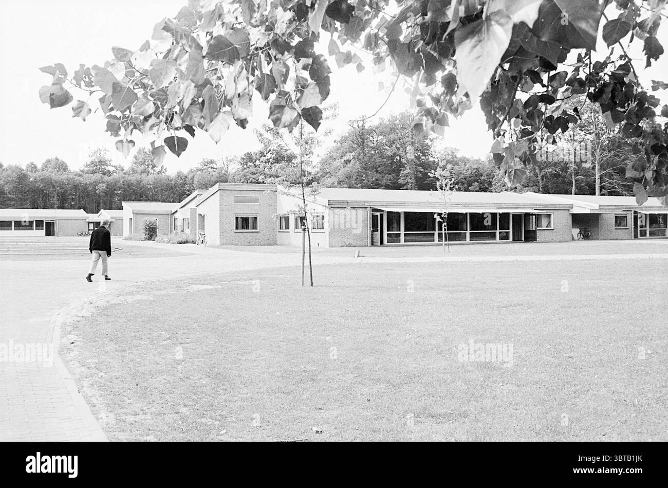 Un complesso scolastico, Whizgle News, Dutch Desk, Paesi Bassi, 1950 - 2000. Questi argomenti sono mostrati nell'immagine. La scena raffigura un ambiente tranquillo all'aperto, caratterizzato da un'ampia area erbosa aperta che si estende in lontananza. In primo piano, una figura solitaria cammina piacevolmente lungo un sentiero curvo. L'individuo sembra essere vestito con abiti scuri, creando un sottile contrasto con i toni più luminosi del paesaggio. Intorno al percorso si trovano diversi edifici moderni e rettangolari con tetti piatti, i cui esterni sono realizzati in mattoni semplici. L'architettura trasmette un des utilitaristico Foto Stock