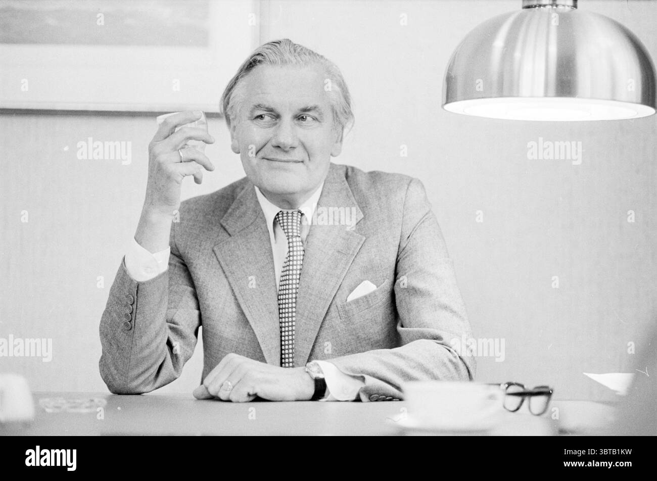 L'Aia, signor Wagner Shell. Persons Companies Den Haag, Whizgle News, Dutch Desk, Paesi Bassi, 1950 - 2000 il 19-10-1982. Questi sono gli elementi dell'immagine. Nella scena, un uomo ben vestito siede a un tavolo, trasuda un senso di fiducia e raffinatezza. Indossa un abito sartoriale grigio chiaro che gli si adatta perfettamente, completato da una classica cravatta a scacchi che aggiunge un tocco di fantasia al suo outfit. I suoi capelli sono ben curati e presentano un aspetto raffinato. L'uomo tiene una bevanda casualmente nella mano destra, le dita si arricciano delicatamente intorno ad essa. La sua espressione è un po' maliziosa, ingegno Foto Stock