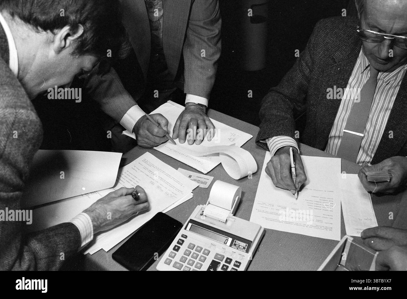LTB fusione WLTO, Whizgle News, Dutch Desk, Paesi Bassi, 1950 - 2000 il 21-05-1991. Questi argomenti vengono visualizzati nell'immagine. Una stanza poco illuminata è piena di un senso di concentrazione e collaborazione. Molte persone sono riunite intorno a un tavolo ricoperto di fogli di carta e di una vecchia calcolatrice. Lo stato d'animo è serio, trasmettendo un senso di urgenza mentre le persone si impegnano in quella che sembra essere una discussione o una negoziazione. In primo piano, sono visibili due mani: Una con una penna posizionata sopra un documento che mostra l'intento, mentre un'altra sta premendo i tasti sulla calcolatrice, eventualmente scricchiolando numeri o tallando il Resul Foto Stock