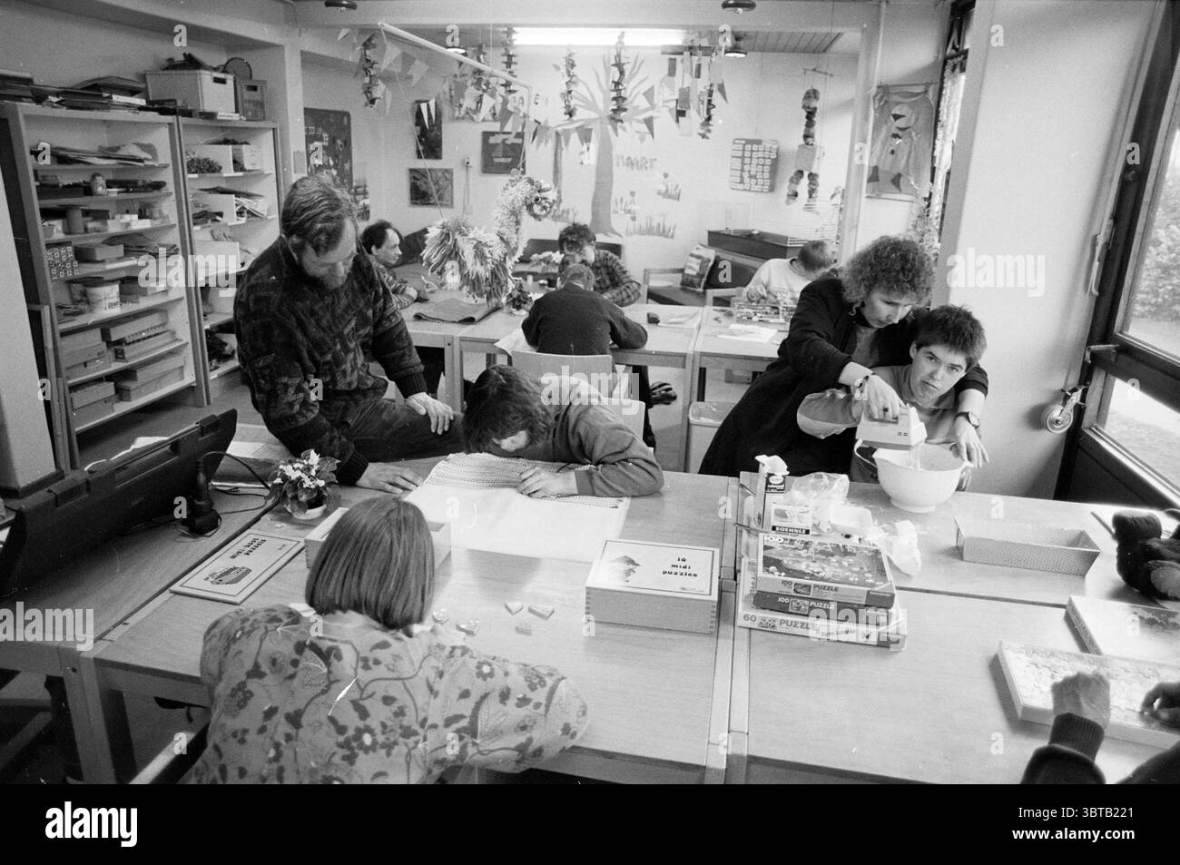 Marie josé Holtgreve e Hans de Berg Cruquiushoeve Persons, Whizgle News, Dutch Desk, Paesi Bassi, 1950 - 2000 il 26-04-1989. L'immagine contiene questi argomenti. La scena si sviluppa in una sala spaziosa e ben illuminata piena di una varietà di tavoli e sedie disposti in modo un po' organizzato. La luce naturale si riversa attraverso le grandi finestre, illuminando le attività e creando un'atmosfera calda e invitante. A un tavolo, diversi individui si stanno impegnando in compiti diversi. Un bambino con un'acconciatura corta si appoggia su una superficie ricoperta di carta e materiali artistici, concentrandosi profondamente sul proprio lavoro Foto Stock