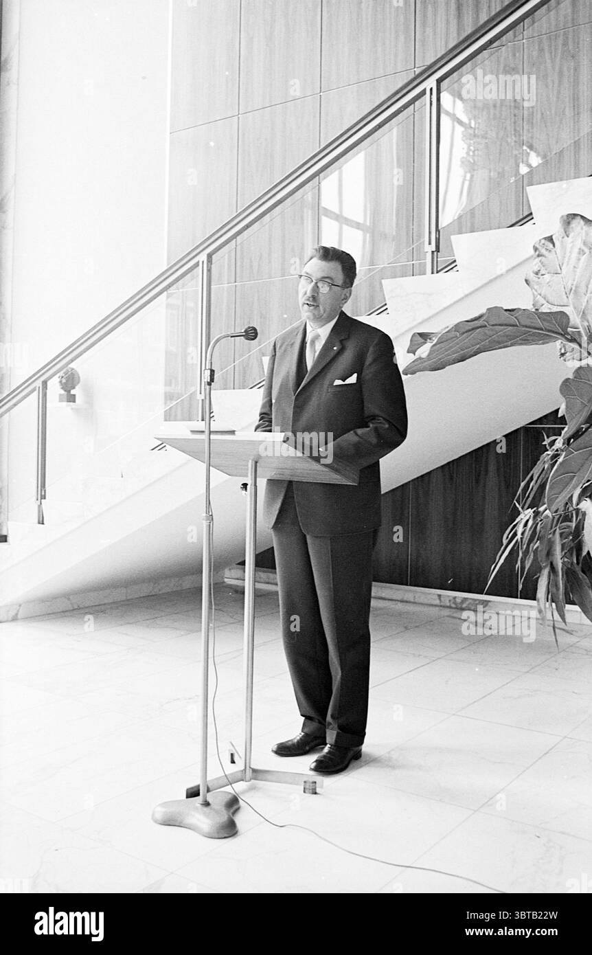1st bus Amsterdam - IJmuiden Noord Zuid Hollandse Vervoersmaatschappij N.V. NZ, Whizgle News, Dutch Desk, Paesi Bassi, 1950 - 2000 il 02-05-1968. L'immagine contiene questi argomenti. La scena raffigura un uomo in piedi dietro un podio di legno, impegnato a rivolgersi a un pubblico. È vestito con un abito scuro, completo di cravatta, che trasuda un'aria di professionalità. I suoi capelli sono ben disegnati, e appare concentrato sul suo discorso, trasmettendo un senso di autorità e fiducia. Lo sfondo presenta interni architettonici moderni, con una scalinata prominente che curva elegantemente verso l'alto. Il wal Foto Stock