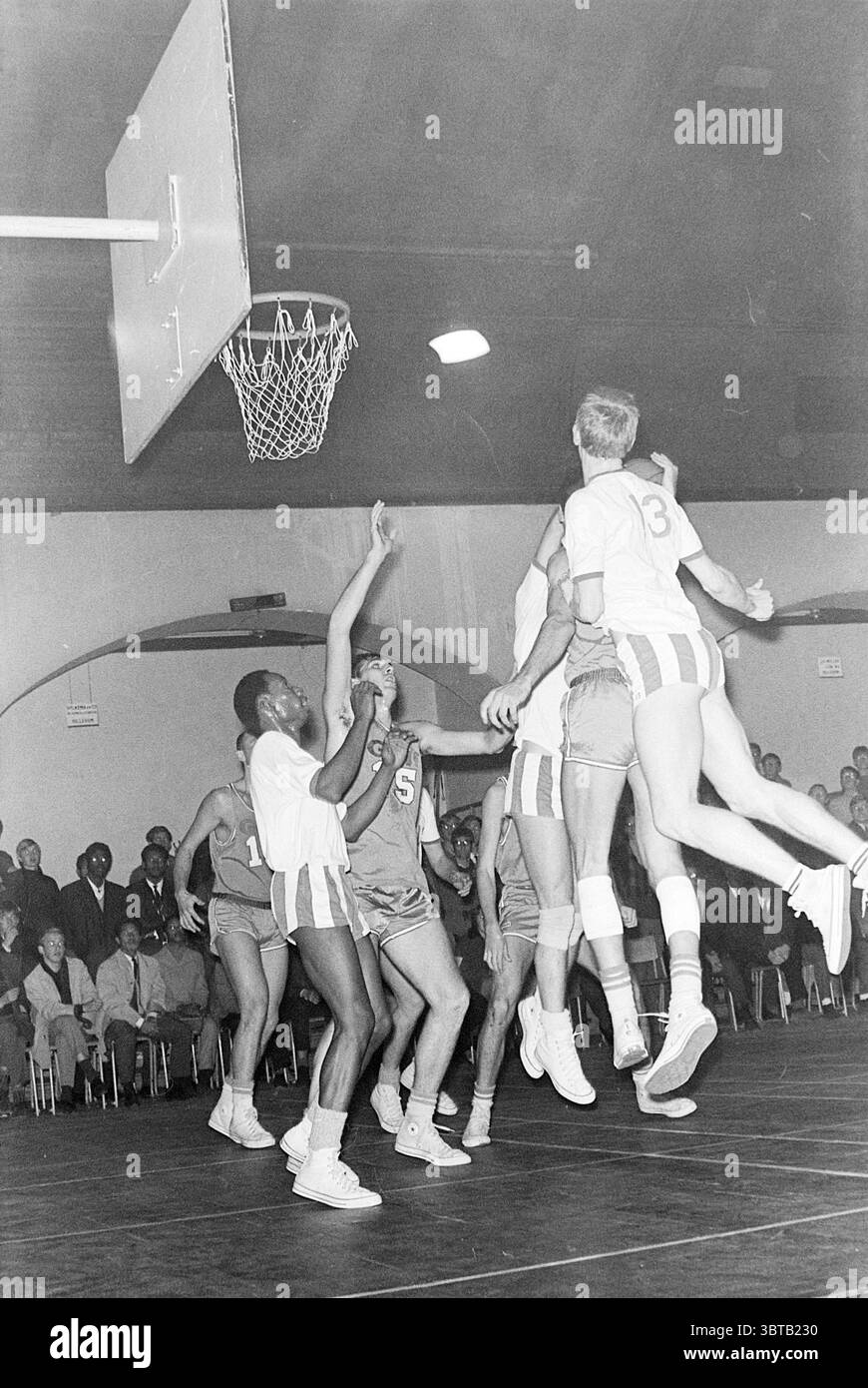 Partita di basket, Whizgle News, Dutch Desk, Paesi Bassi, 1950 - 2000 su 11-1967. Questi argomenti sono mostrati nell'immagine. La scena cattura un momento dinamico in una partita di basket, mostrando l'energia e l'intensità di uno sport competitivo. In primo piano, i giocatori in uniforme atletica sono impegnati in un gioco feroce, con un giocatore che salta in alto per fare un tiro verso il cerchio. Questo atleta si distingue in modo evidente contro gli altri, indossando una maglia di colore chiaro con il numero "13" sul retro, che contrasta con le tonalità più scure degli abiti dei suoi compagni di squadra. Gli altri giocatori sono posizionati a Foto Stock