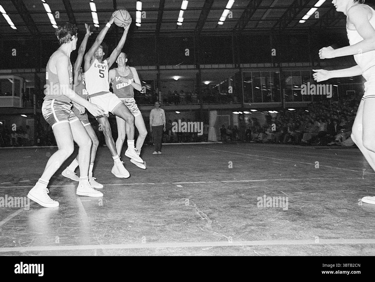 Basket, Whizgle News, Dutch Desk, Paesi Bassi, 1950 - 2000 il 05-1971. Questi sono gli elementi dell'immagine. La scena cattura un momento dinamico in una partita di basket. In primo piano, un giocatore con una maglia scura con il numero "5" salta in aria, raggiungendo il pallacanestro appena sopra la sua testa, mostrando un'espressione decisa. La sua struttura atletica enfatizza lo sforzo del salto, con gli arti allungati in modo drastico. Intorno a lui ci sono due avversari, entrambi con maglie di colore più leggero, che cercano di difendersi dal suo tiro. Un giocatore ha le braccia sollevate, tentando di bloccare la palla Foto Stock