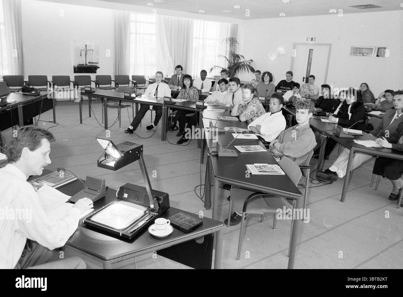 Studenti di inglese presso la camera di commercio di Haarlem Nederland, Whizgle News, Dutch Desk, Paesi Bassi, 1950 - 2000 il 14-04-1991. Questi argomenti sono mostrati nell'immagine. La scena raffigura una sala riunioni piena di persone sedute intorno a pochi grandi tavoli disposti a U. L'ambiente è professionale, caratterizzato da un'estetica moderna con linee pulite. In prima linea, un altoparlante, vestito con una camicia di colore chiaro con colletto, è posizionato su uno dei tavoli, indirizzando l'attenzione su un proiettore o su una fotocamera per documenti. L'apparecchiatura emette un debole bagliore, illuminato contro l'altro ne Foto Stock