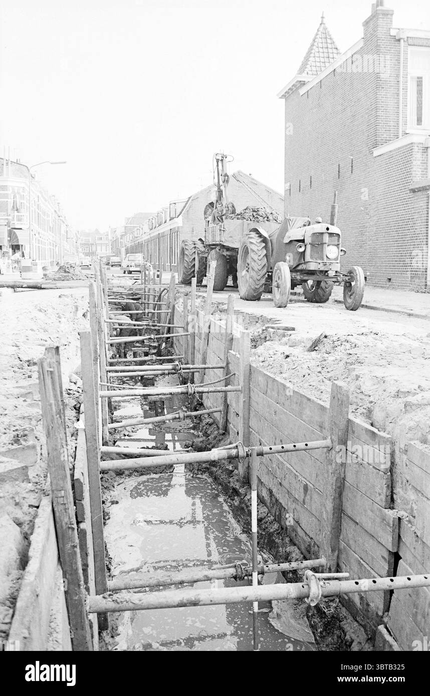 Fognatura - Kenaupark Kleverparkstraat Spanjaardslaan Iordenstraat Sewerage Haarlem Spanjaardslaan Paesi Bassi, Whizgle News, Dutch Desk, Paesi Bassi, 1950 - 2000 il 16-09-1965. L'immagine mostra questi argomenti. La scena raffigura un cantiere in un ambiente urbano, caratterizzato da una trincea che si trova in primo piano. La trincea è circondata da supporti in legno, che indicano i lavori infrastrutturali in corso, probabilmente relativi a linee idrauliche o di pubblica utilità. Le pareti della trincea sono rinforzate con travi di legno, che mostrano segni di umidità dalla terra. Sullo sfondo, a Foto Stock