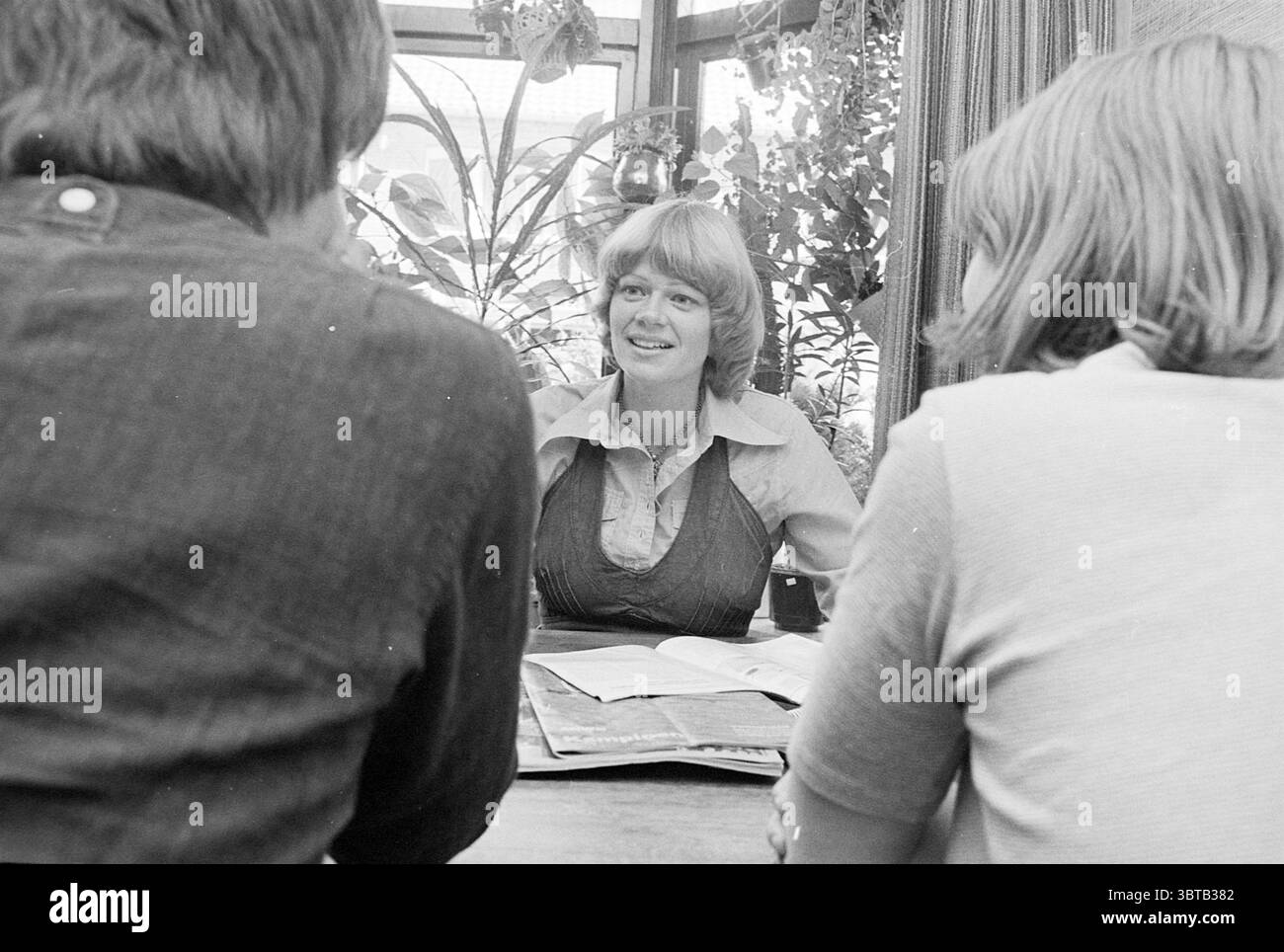 Portrait Woman, Whizgle News, Dutch Desk, Paesi Bassi, 1950 - 2000 nel 1977. Questi sono gli elementi dell'immagine. In un intimo ambiente interno, tre persone sono sedute attorno ad un tavolo di legno, creando un'atmosfera intima. La donna in primo piano ha un sorriso caldo e un comportamento coinvolgente, indossando una camicetta di colore chiaro che contrasta con i toni più scuri dell'abbigliamento dei suoi compagni. I suoi capelli sono modellati in modo da suggerire un look rilassato e casual. Le due persone di fronte a lei si girano leggermente di lato, mostrando il loro interesse. Uno ha una camicia scura, che aggiunge profondità alla scena, mentre l'altro Foto Stock