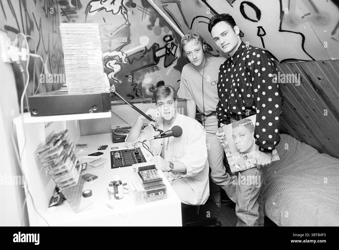 Music in the ether 3 gentlemen at mixing console, Whizgle News, Dutch Desk, Paesi Bassi, 1950 - 2000 on 09-1989. L'immagine contiene questi argomenti. La scena cattura uno spazio di registrazione accogliente e informale con un'atmosfera eclettica. Le pareti sono adornate da vivaci graffiti, che mostrano un mix di colori e forme astratte che aggiungono un tocco urbano. In primo piano, tre persone occupano la piccola stanza. Una persona è seduta a una scrivania, concentrandosi su una console di miscelazione dotata di varie manopole e pulsanti. Il suo abbigliamento è casual e presenta una camicia oversize di colore chiaro. Accanto a lui, altri due Foto Stock