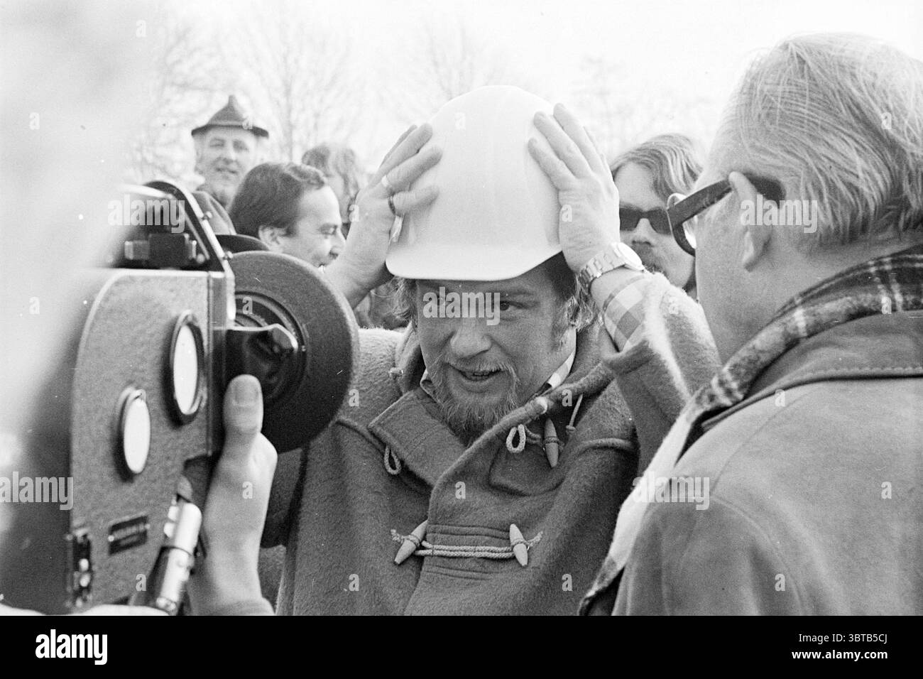 Primo mucchio di case di diritto immobiliare Haarlemmerliede dal Segretario di Stato Schaeffer primo mucchio, Whizgle News, Dutch Desk, Paesi Bassi, 1950 - 2000 il 10-02-1975. Questi sono gli elementi dell'immagine. La scena è ambientata all'aperto, probabilmente durante un evento significativo, forse un progetto di costruzione o di innovazione. Un gruppo di persone è riunito, suggerendo un'atmosfera festosa o memorabile. Il primo piano presenta un individuo che mette un elmetto bianco sulla testa, a significare un senso di responsabilità o partecipazione a un'attività di costruzione. Questa persona ha un'espressione concentrata, suggerisce Foto Stock