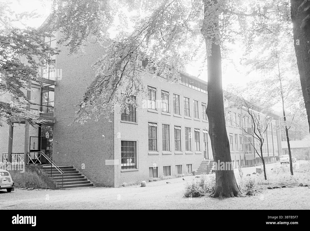 Lavigerie College Santpoort Schools, Whizgle News, Dutch Desk, Paesi Bassi, 1950 - 2000 il 27-05-1968. L'immagine include questi argomenti. La scena cattura un momento sereno e un po' nostalgico, caratterizzato da un grande edificio in mattoni che suggerisce uno stile storico o istituzionale. La struttura è caratterizzata dalle sue finestre rettangolari, molte delle quali si estendono dal pavimento al soffitto, consentendo l'ingresso di un'ampia luce naturale all'interno. La muratura ha un tono tenue, trasuda un senso di permanenza e stabilità. A sinistra, una serie di gradini conduce all'ingresso, incorniciato da un rai di legno Foto Stock