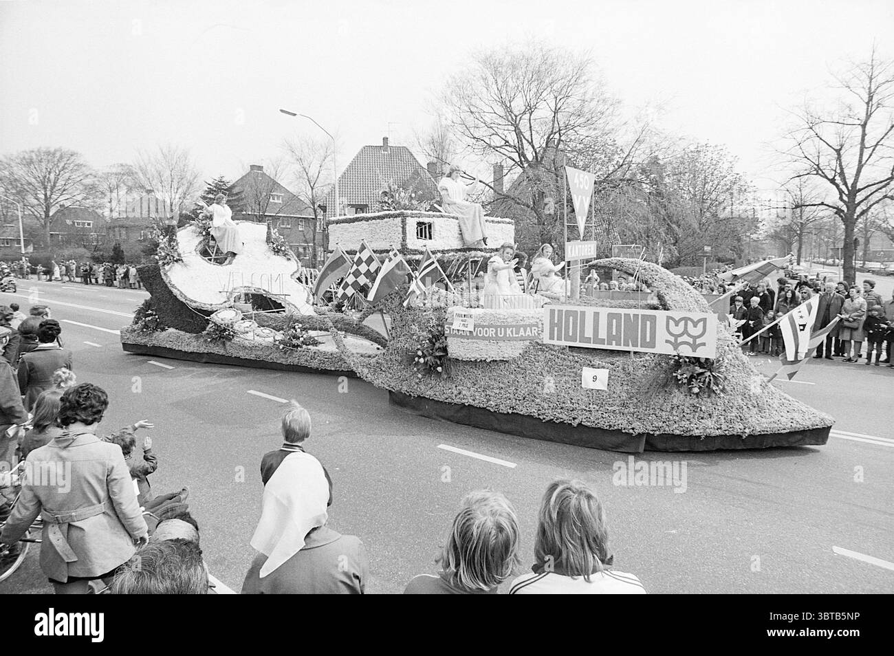 Flower Parade.., Whizgle News, Dutch Desk, Paesi Bassi, 1950 - 2000. L'immagine contiene questi argomenti. In questa vivace scena, una sfilata festosa si svolge lungo un'ampia strada asfaltata. Il punto focale è un sontuoso galleggiante adornato da fiori e vegetazione vibranti, disposto ad arte per creare disegni intricati. Il galleggiante appare festoso e grandioso, somiglia a una nave, con curve ampie e grandi elementi decorativi che imitano le onde. Intorno al galleggiante, le persone fiancheggiano le strade, mostrando un mix di emozioni, dall'emozione alla gioia. La folla è variegata, con adulti e bambini, alcuni dei quali battono bandiera Foto Stock
