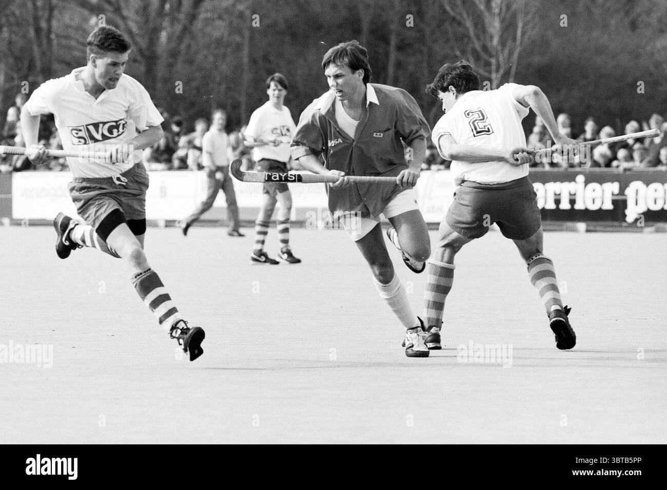 Hockey Bloemendaal - Tilburg, Whizgle News, Dutch Desk, Paesi Bassi, 1950 - 2000 il 16-03-1991. L'immagine mostra questi argomenti. La scena cattura un momento intenso su un campo di hockey, in cui tre atleti sono attivamente impegnati in una partita. Due giocatori in maglie bianche gareggiano contro uno in una vivace maglia arancione. L'atleta in arancione sembra manovrare la palla abilmente, con un'espressione concentrata, mentre il giocatore alla sua sinistra sembra cercare di intercettare il gioco. I due giocatori in bianco mostrano numeri distinti: Uno mostra in modo prominente il numero 2, mentre l'altro ha una m. Foto Stock