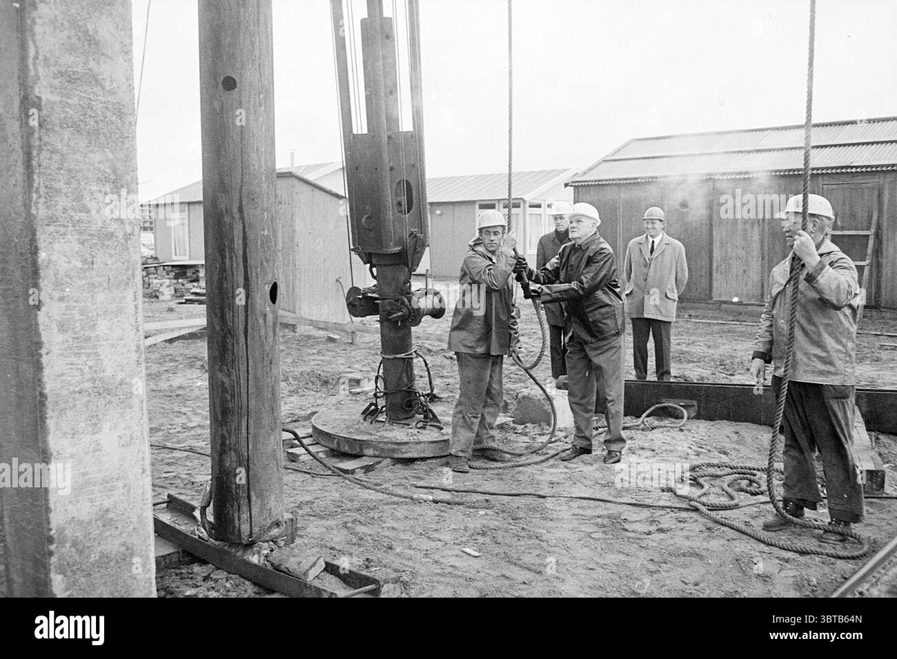 Primo mucchio di case Meerwijk primo mucchio Meerwijk, Whizgle News, Dutch Desk, Paesi Bassi, 1950 - 2000 il 17-10-1968. Questi sono gli argomenti nell'immagine. In un cantiere, quattro persone sono impegnate in un'attività che sembra coinvolgere macchinari. Lo sfondo presenta diverse basse strutture in legno, probabilmente strutture di stoccaggio o di lavoro, e il terreno è coperto da un mix di sporco e ghiaia, suggerendo un'area di costruzione attiva. La scena è dominata da grandi attrezzature industriali, in particolare da un imponente carro di perforazione, che ha una struttura cilindrica nera con tubi flessibili Foto Stock