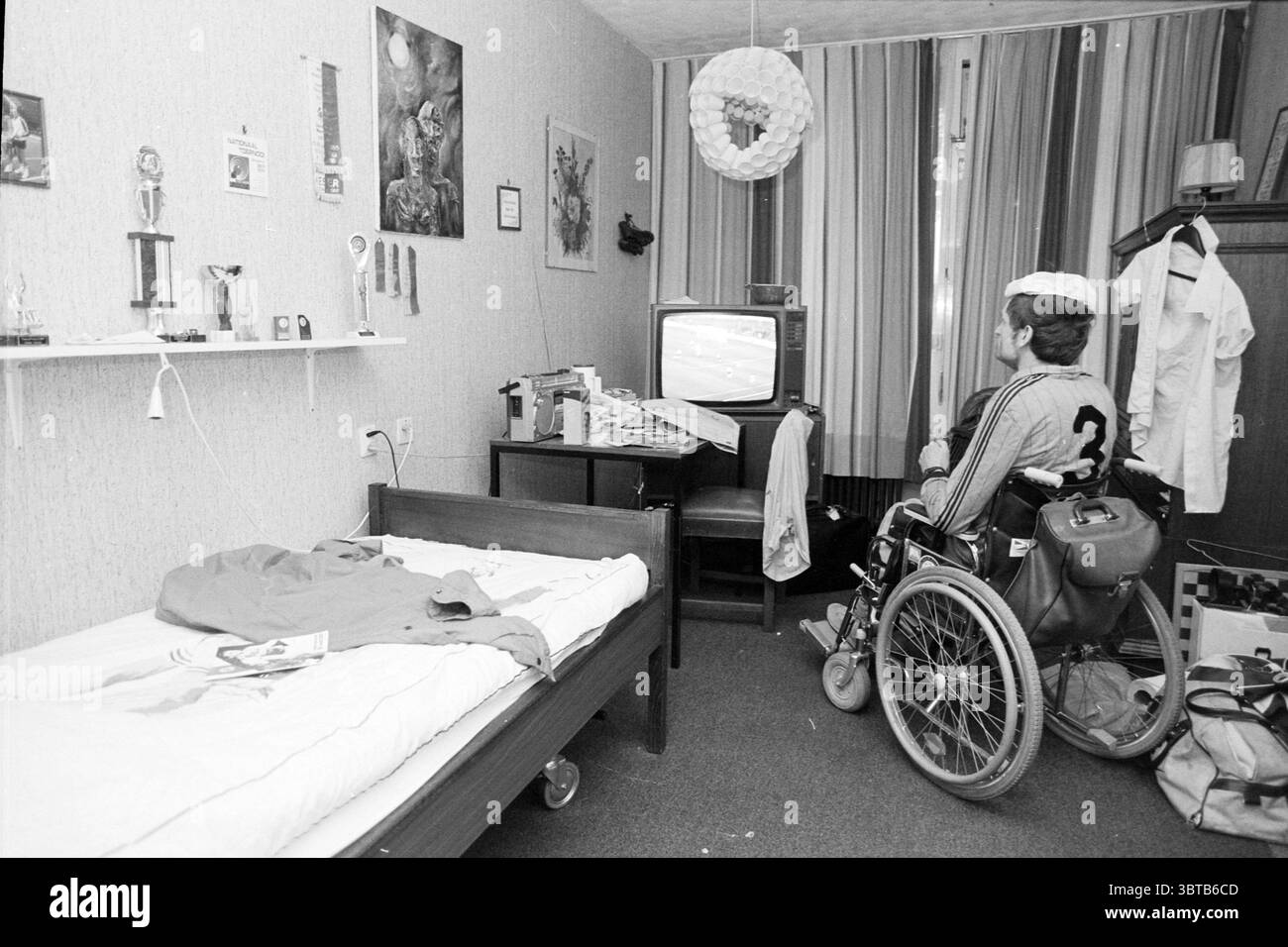 Guarda le semifinali Paesi Bassi - Germania 3 volte calcio, Whizgle News, Dutch Desk, Paesi Bassi, 1950 - 2000 il 21-06-1988. Questi argomenti sono mostrati nell'immagine. La scena presenta una stanza moderatamente arredata caratterizzata da un mix di oggetti personali e oggetti di uso quotidiano. Da un lato, c'è un letto in stile ospedale, le sue lenzuola non fatte e alcuni vestiti sparsi che suggeriscono uno spazio vissuto. Una sedia a rotelle suggerisce le esigenze di mobilità dell’occupante. Sullo sfondo, una piccola scrivania è piena di oggetti vari, tra cui libri e forse alcuni documenti, insieme a un televisore d'epoca Foto Stock