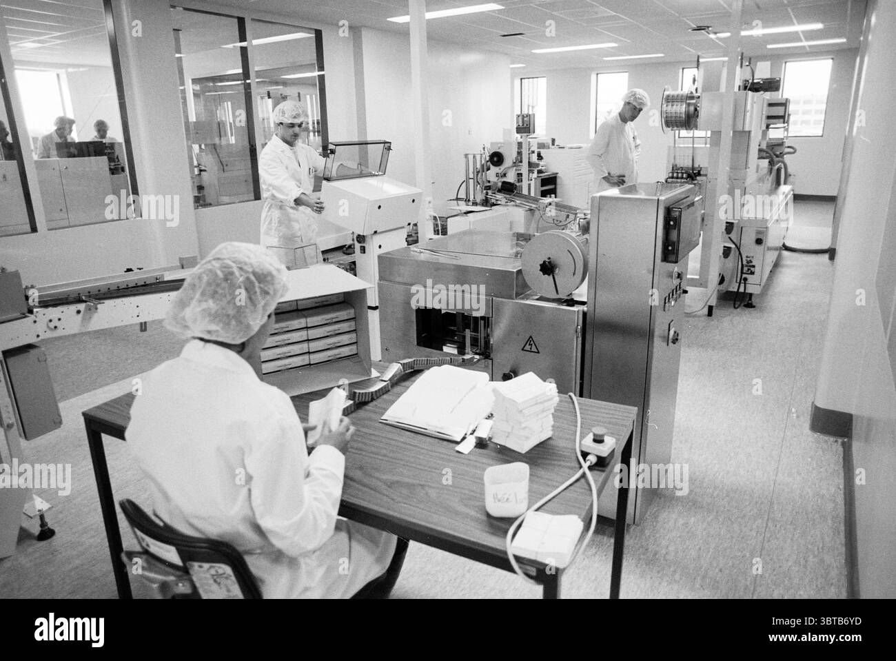 Alcune persone presso macchine per il confezionamento farmaceutico di Pharma Chemie, Whizgle News, Dutch Desk, Paesi Bassi, 1950 - 2000 il 16-05-1990. L'immagine include questi argomenti. La scena raffigura un laboratorio sterile e ben illuminato o un'area di produzione incentrata su un processo meticoloso. Lo spazio è dominato da una gamma di macchinari e attrezzature industriali progettati per la precisione e l'efficienza. In primo piano, una persona vestita con un camice da laboratorio bianco siede su un tavolo di legno, impegnata nella manipolazione di piccoli imballaggi o materiali, eventualmente nell'esecuzione di un'ispezione o nella preparazione di articoli per un'ulteriore lavorazione. T Foto Stock