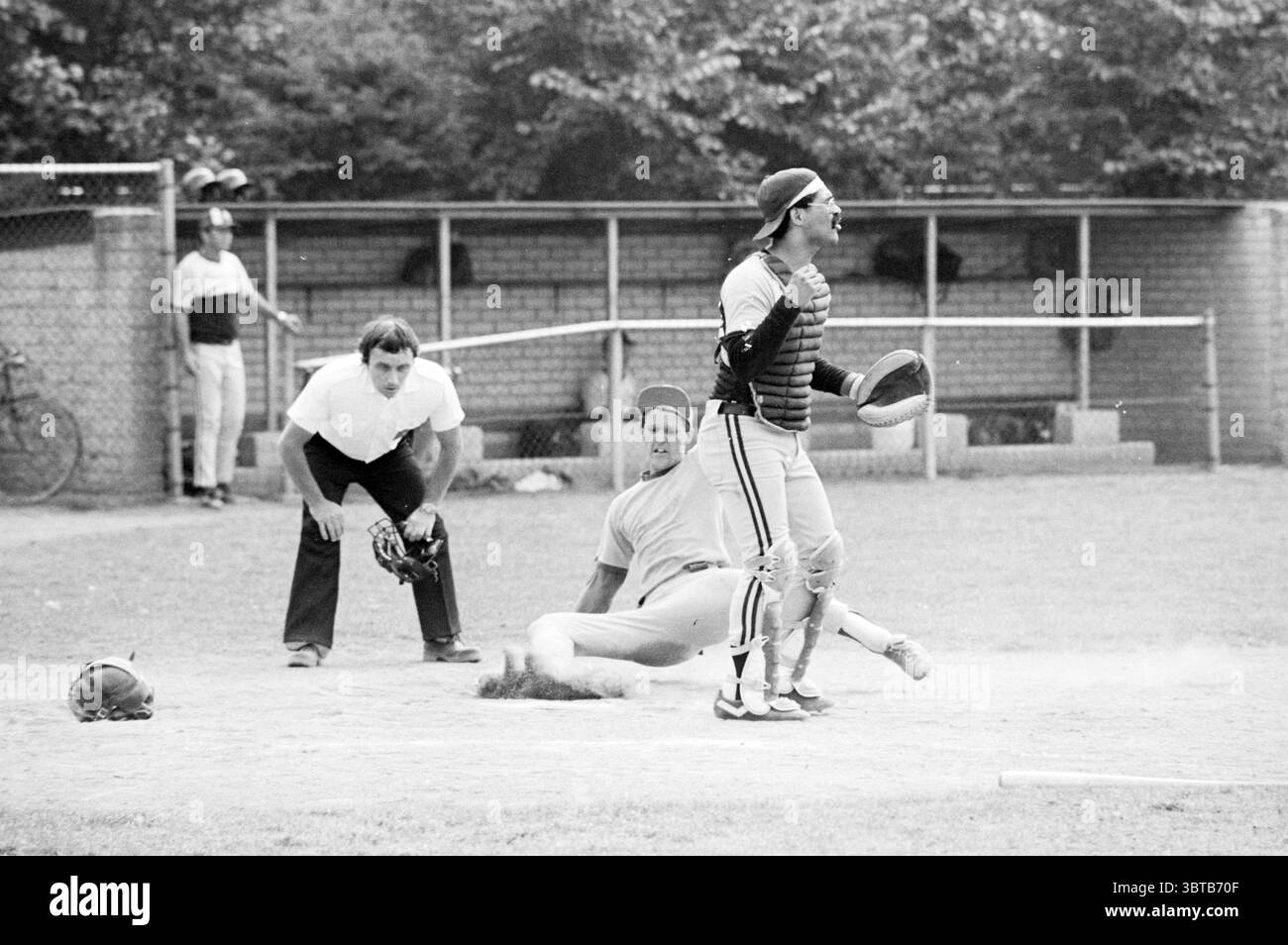 Rooswijk- Black Lions Baseball Rooswijk, Whizgle News, Dutch Desk, Paesi Bassi, 1950 - 2000 il 31-07-1982. L'immagine mostra questi argomenti. La scena cattura un momento dinamico su un campo da baseball erboso. In primo piano, un giocatore che scivola nella piastra di casa è il punto focale, raffigurato a metà movimento con un'espressione di determinazione. È rivestito con un'uniforme di colore chiaro con strisce scure, e il suo corpo è angolato mentre si allunga verso il piatto. Dietro di lui, un ricevitore accovacciato, indossando un casco nero e una protezione del petto. La sua postura è assertiva, indicando che è pronto a ricevere l'incom Foto Stock