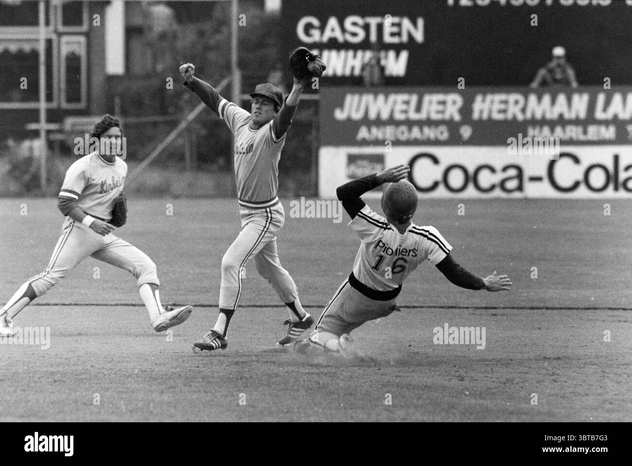 Kinheim - Hoofddorp Baseball Kinheim, Whizgle News, Dutch Desk, Paesi Bassi, 1950 - 2000 il 02-07-1983. Questi argomenti sono mostrati nell'immagine. In una dinamica scena all'aperto, due giocatori di squadre di baseball avversarie vengono catturati in azione su un campo erboso. Un giocatore, indossando un'uniforme grigio chiaro con accenti arancioni, sta in piedi sul campo, con il guanto sollevato in alto, segnalando un momento cruciale, probabilmente una palla catturata o un gioco imminente in una base. La sua postura riflette determinazione e concentrazione. Alla sua sinistra, un giocatore dell'altra squadra è a metà corsa verso la base, il suo corpo basso verso il groun Foto Stock