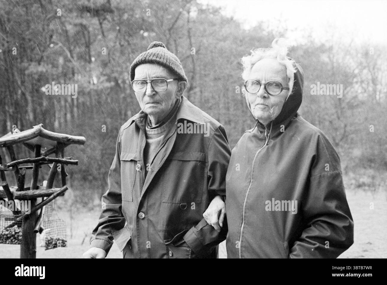 Jan Strijbos con moglie Nature People, Whizgle News, Dutch Desk, Paesi Bassi, 1950 - 2000 il 09-03-1981. Questi sono gli elementi dell'immagine. La scena cattura un momento cupo con una coppia anziana che si trova a stretto contatto, trasudando un senso di compagnia. L'uomo che indossa una giacca pratica e scura e un cappello in maglia ha un'espressione seria, con gli occhiali che accentuano il suo sguardo attento. Accanto a lui, la donna è vestita con un cappotto con cappuccio, i suoi occhiali riposano vicino alle sue prominenti zigomi. La sua espressione è altrettanto seria, riflettendo una comprensione condivisa e una profondità di esperienza. Foto Stock