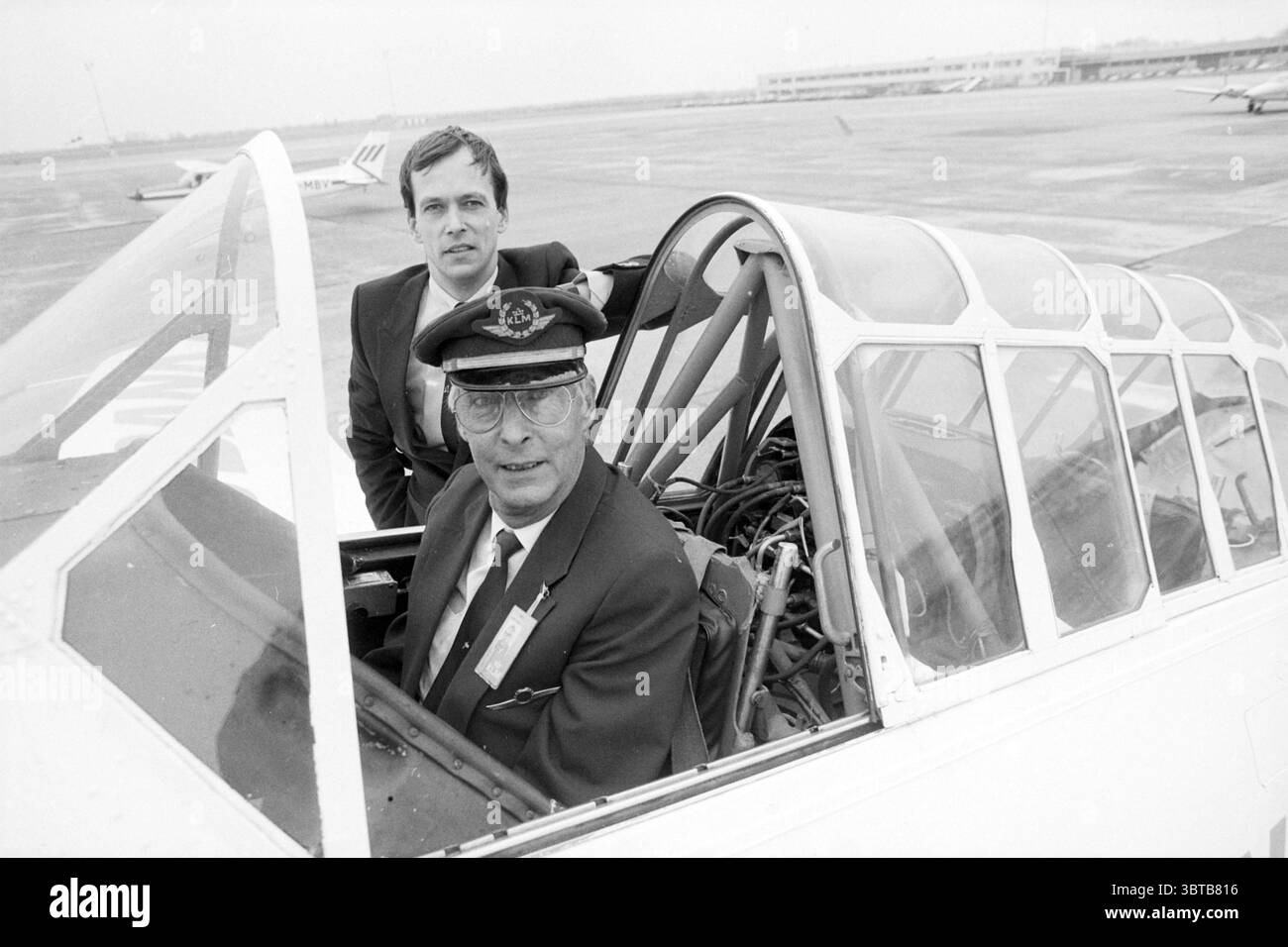 KLM capitano della Geest and Son a causa dell'ultimo volo KLM, Whizgle News, Dutch Desk, Paesi Bassi, 1950 - 2000 il 20-02-1987. L'immagine include questi argomenti. In un vivace ambiente aeroportuale, due persone occupano la cabina di pilotaggio di un aereo d'epoca. La prima persona, in primo piano, indossa un'uniforme ufficiale da pilota completa di cappello. La sua espressione è fiduciosa, riflettendo orgoglio e autorità. È seduto al posto del pilota, circondato dall'intricato cruscotto, con numerosi quadranti e comandi che suggeriscono la complessità operativa dell'aereo. Dietro di lui c'è un secondo uomo, un po' beh Foto Stock