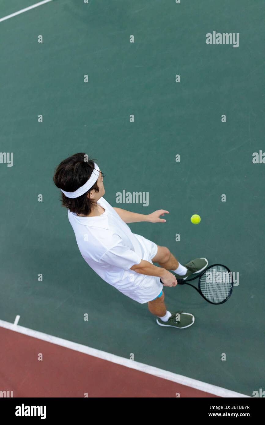 Tennista asiatico maschile in bianco che lancia palla gialla e racchetta avvincente sul campo con linee Foto Stock