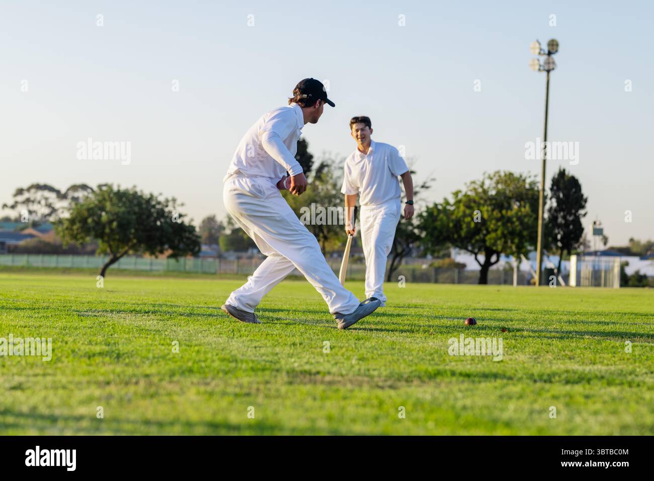 Due compagni di squadra maschili in uniforme bianca si muovono per fermare la palla di cricket in un terreno periferico con la mazza Foto Stock