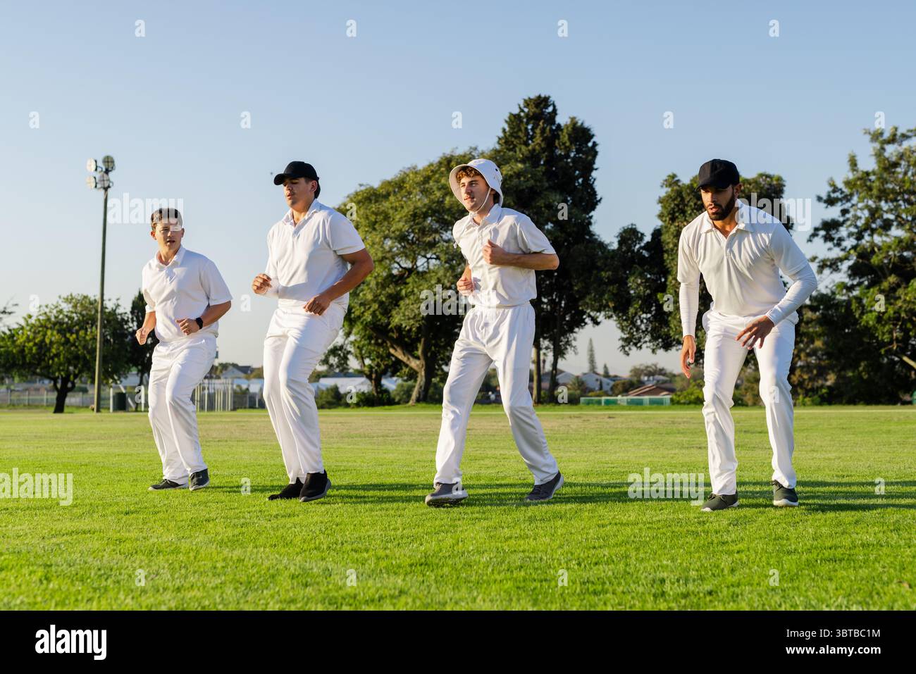 Diversi compagni di squadra maschili accovacciati sul campo di cricket di periferia indossando uniformi bianche sotto il cielo soleggiato Foto Stock
