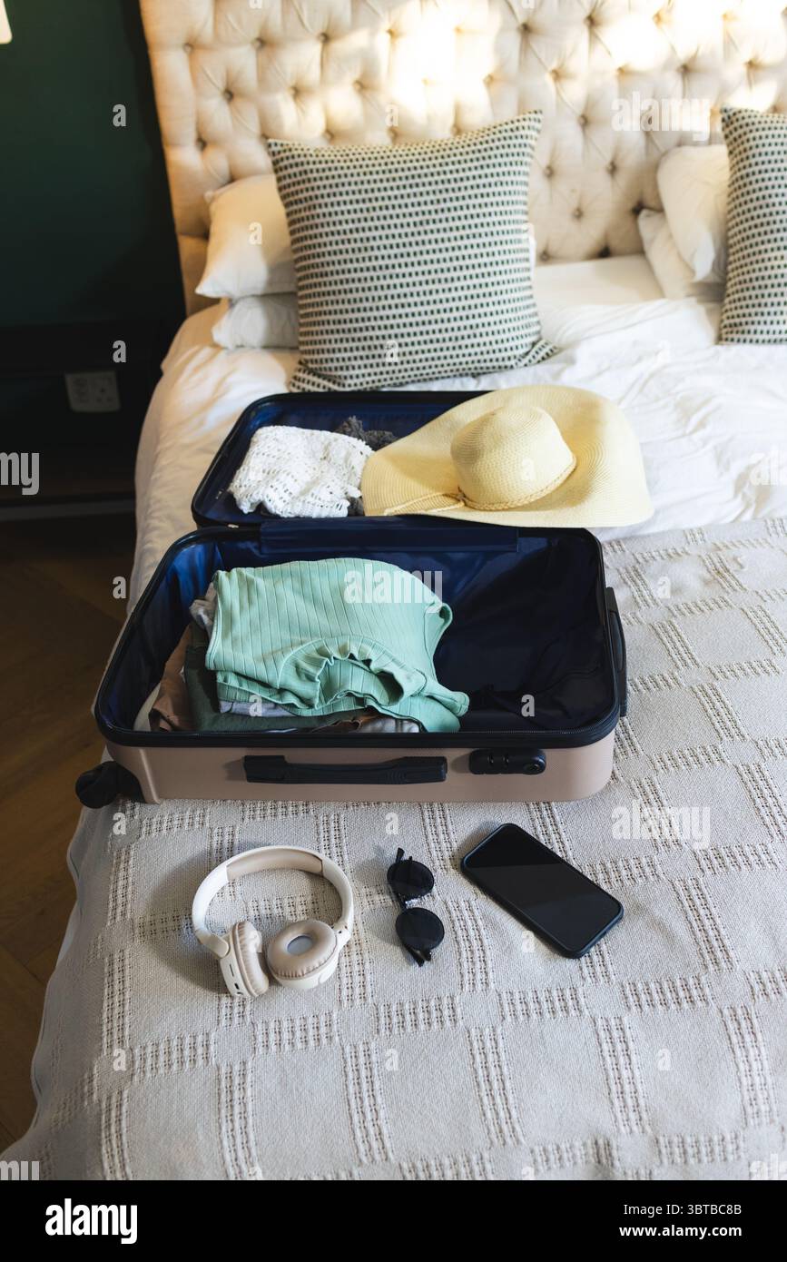 La valigia open tan si appoggia sul letto e contiene una crochet top e un cappello da sole, cuffie e occhiali da sole Foto Stock